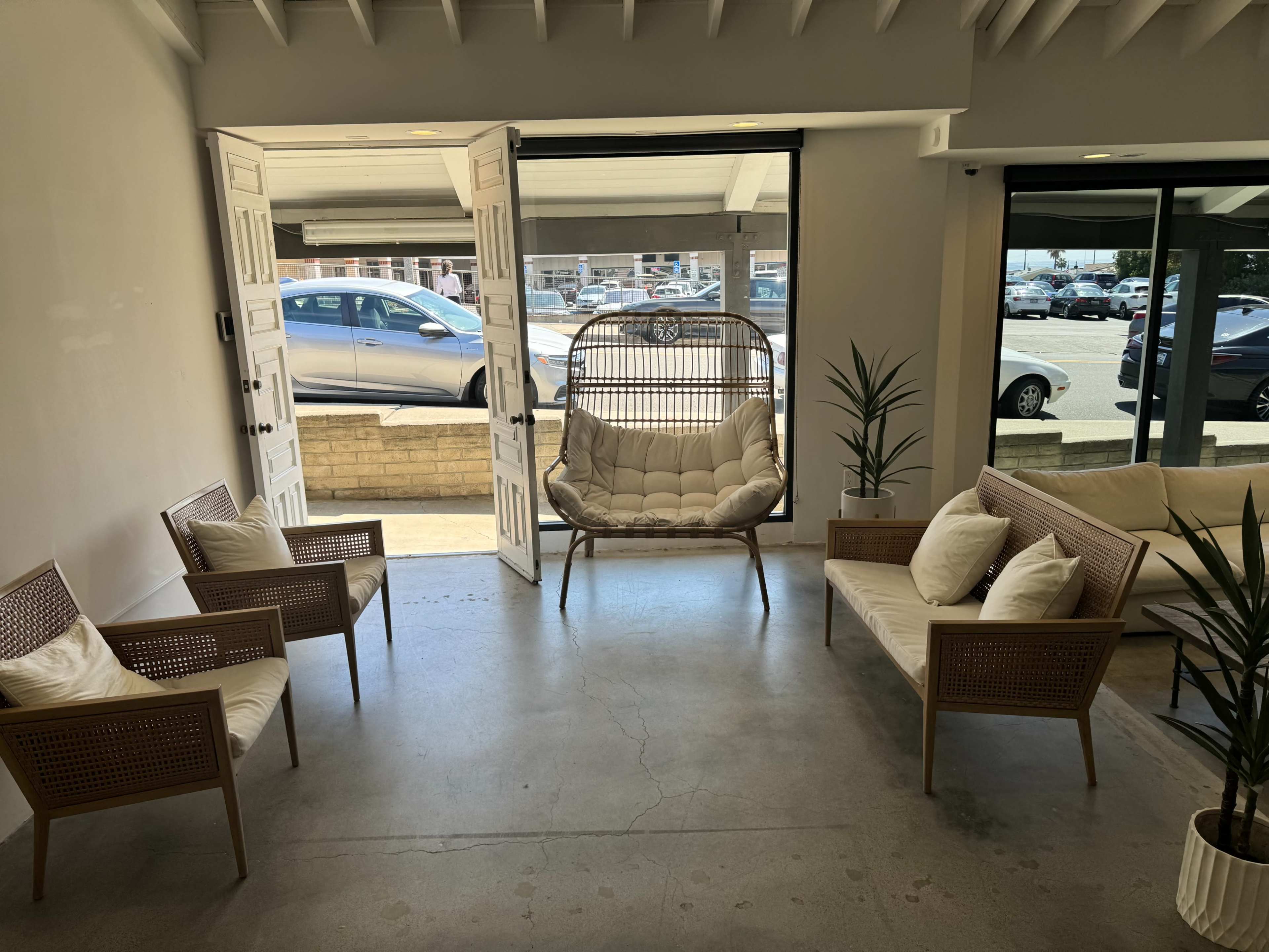 A modern lounge area with several chairs and a hanging chair, facing open double doors that lead to a parking lot.