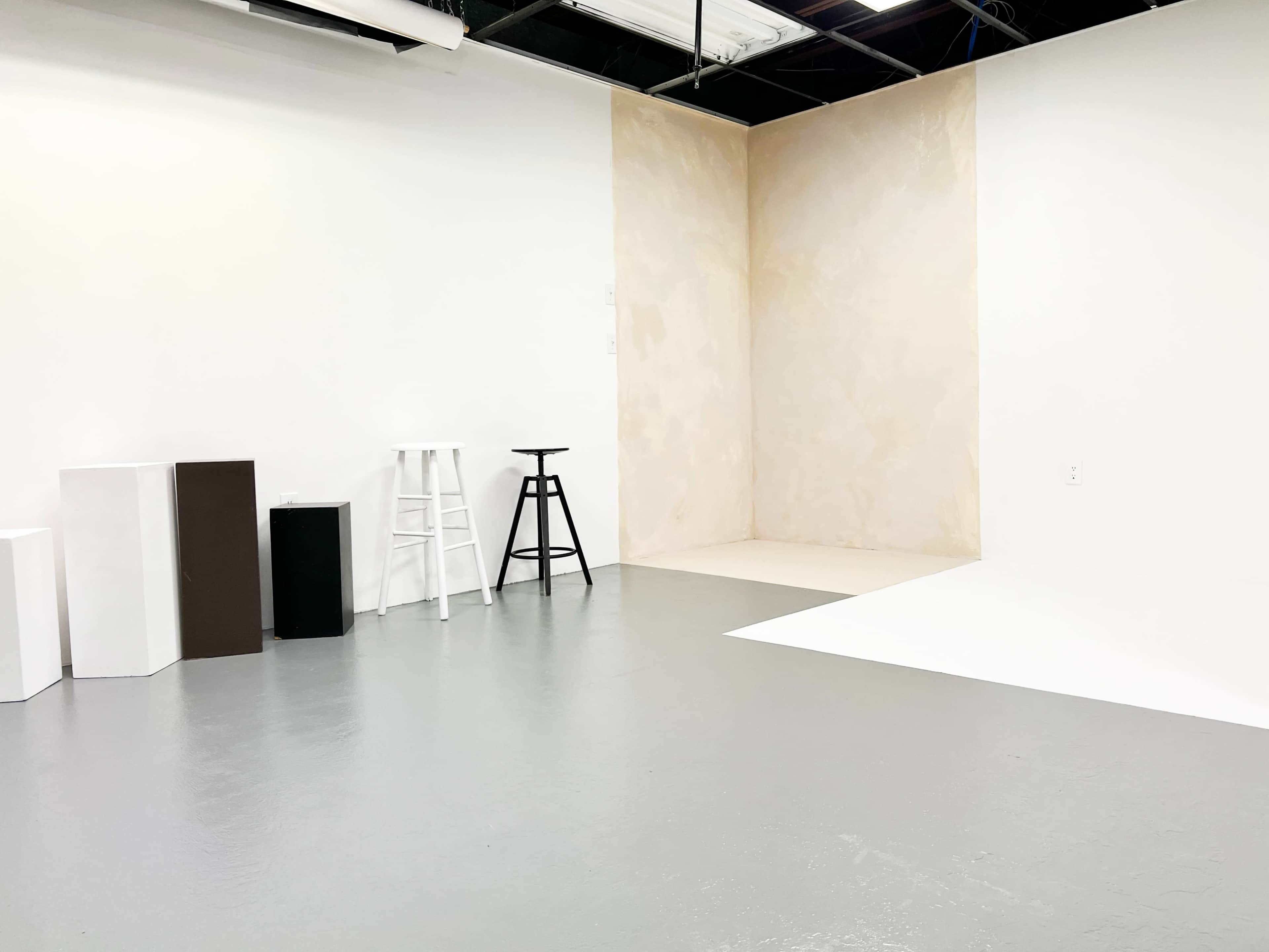 The image depicts a minimalistic studio space with a light-colored wall, variously shaped stands, and a smooth gray floor.