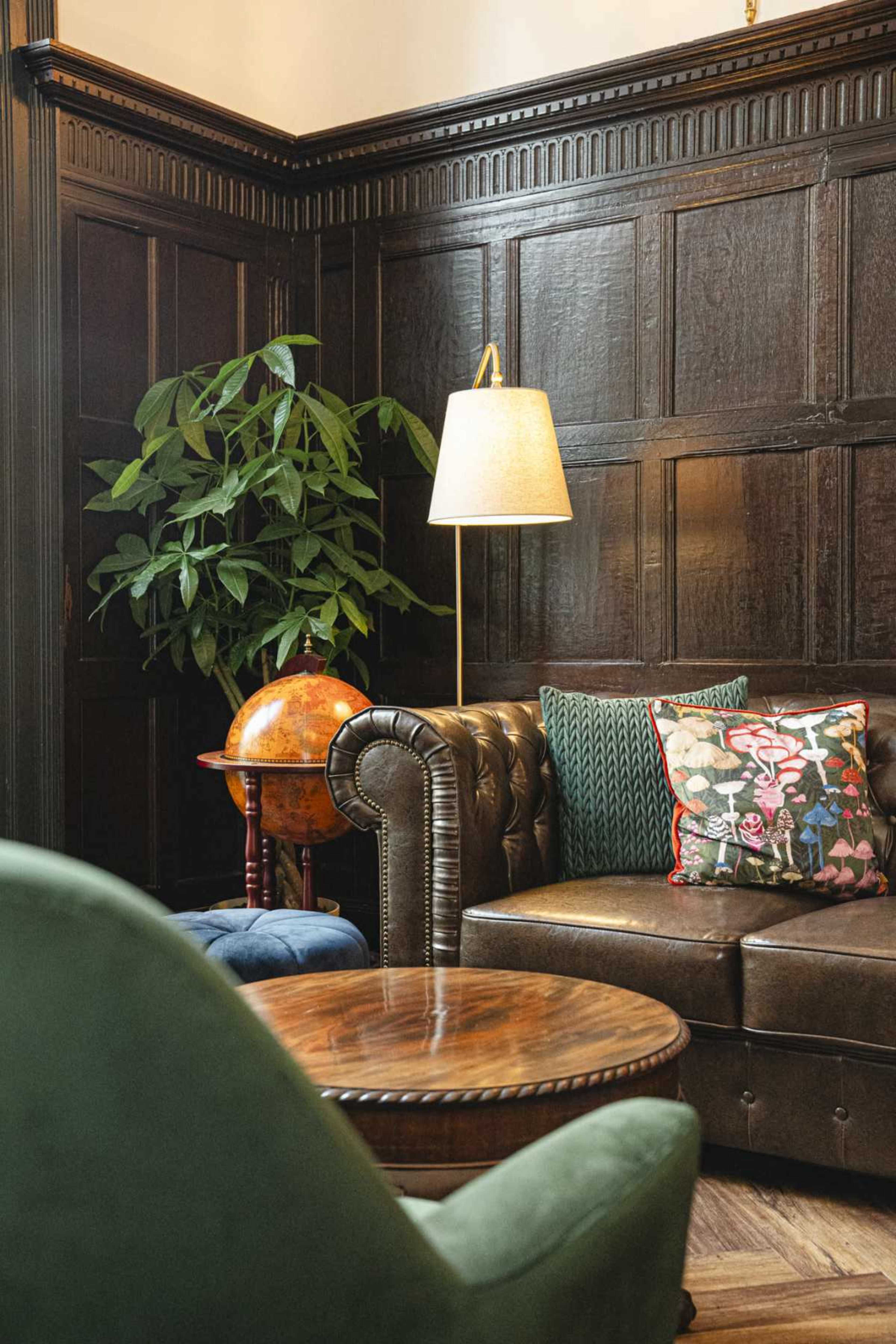 The image shows a cozy seating area with a brown leather couch, a round wooden coffee table, and a green accent chair, accompanied by a floor lamp and a globe on a stand.