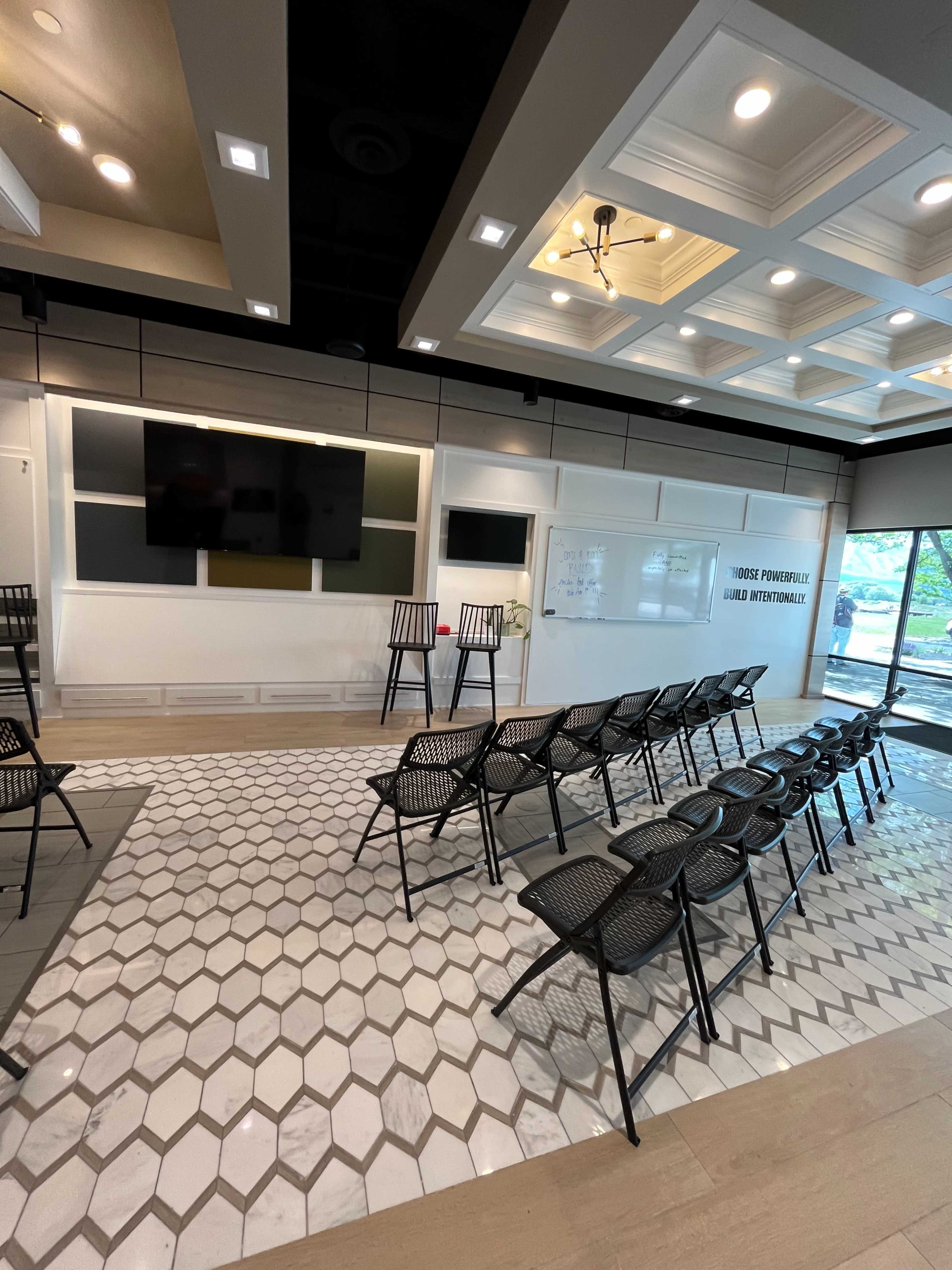 The image shows a modern meeting room with a seating area of black chairs arranged in rows, a large whiteboard, and two mounted screens on the wall.