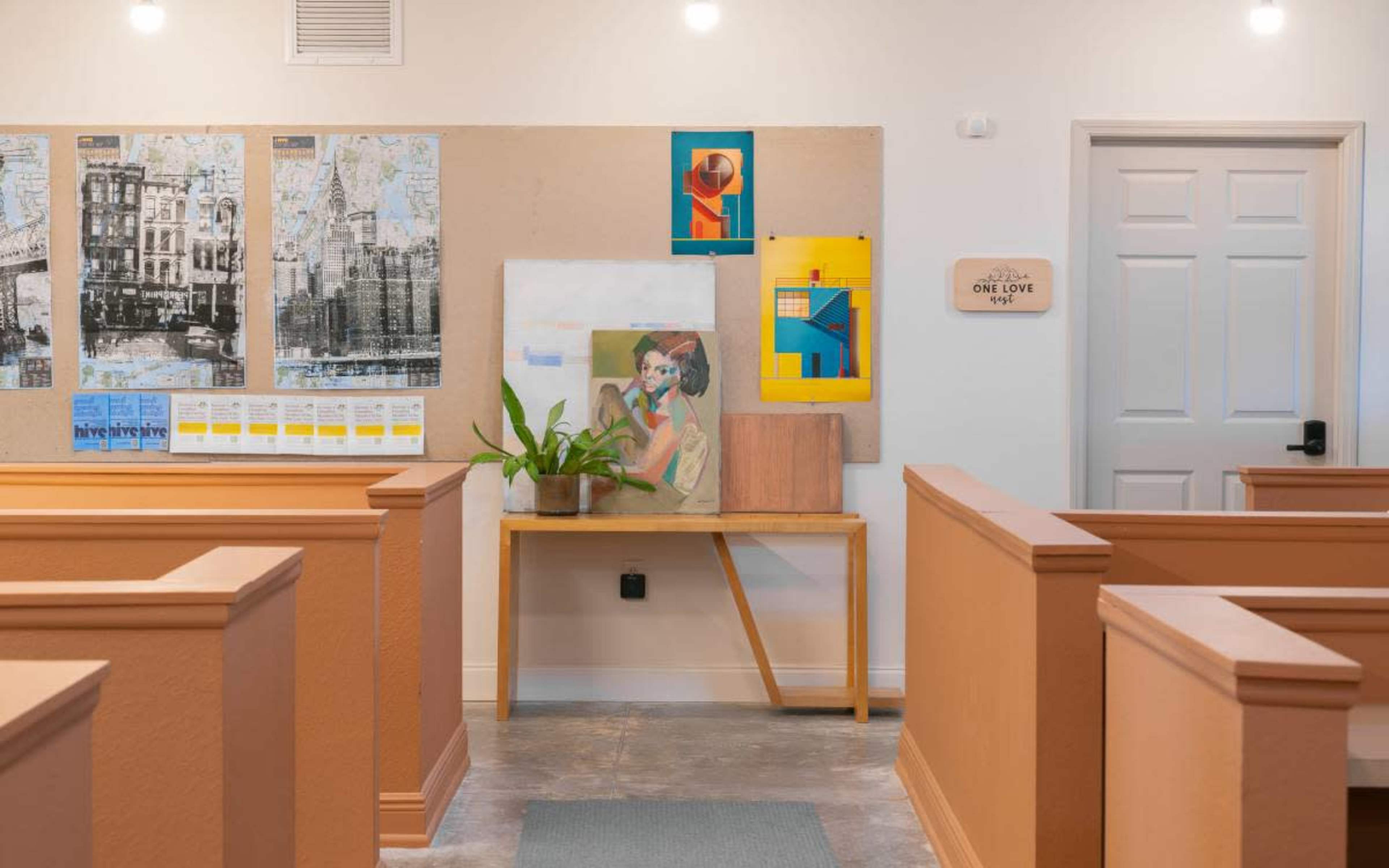 The image shows a corridor with orange walls, wooden benches on either side, and an art display featuring a painting and various posters mounted on a bulletin board.