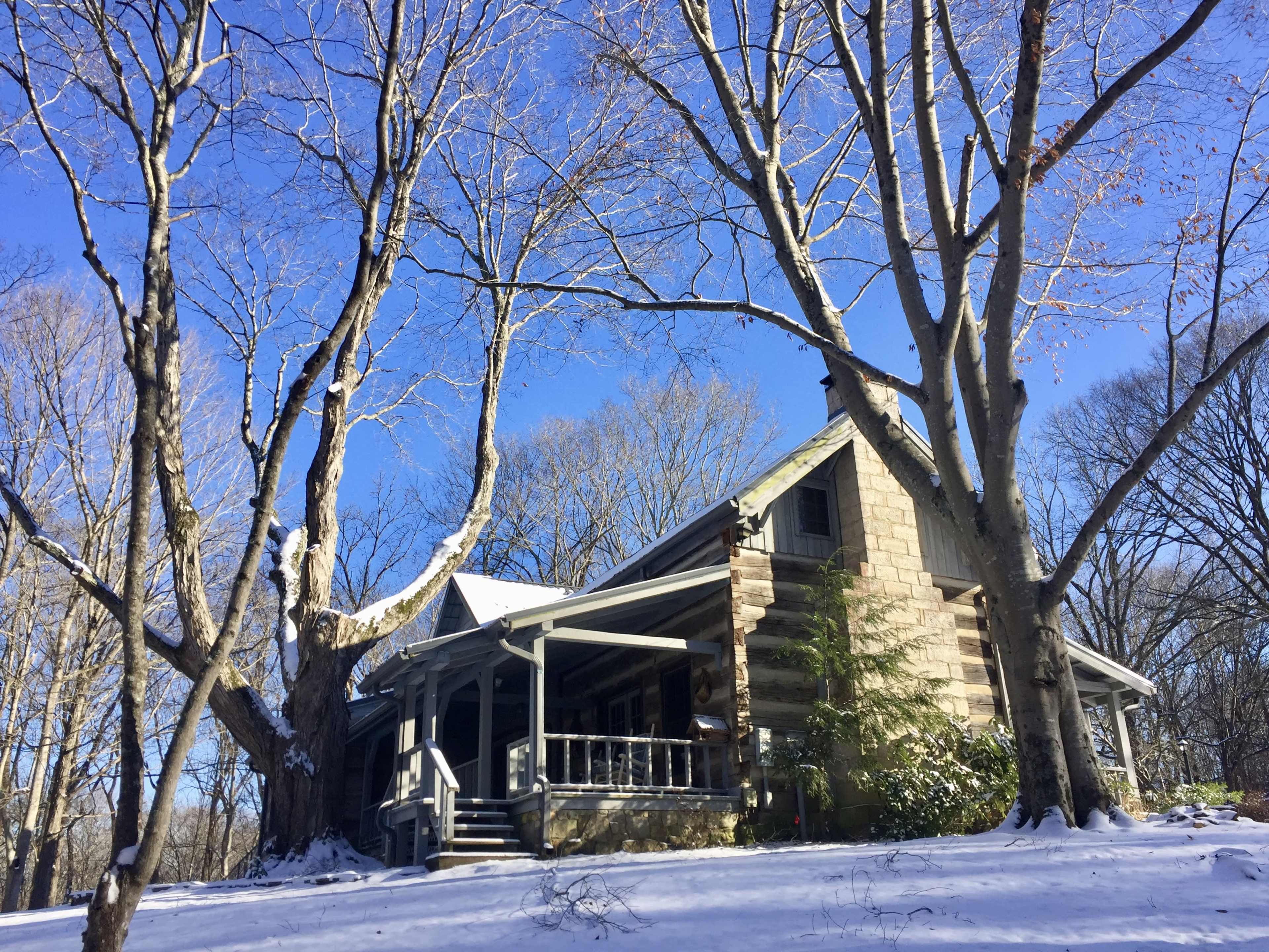 A stone house with a porch is surrounded by bare trees and a blanket of snow under a clear blue sky.