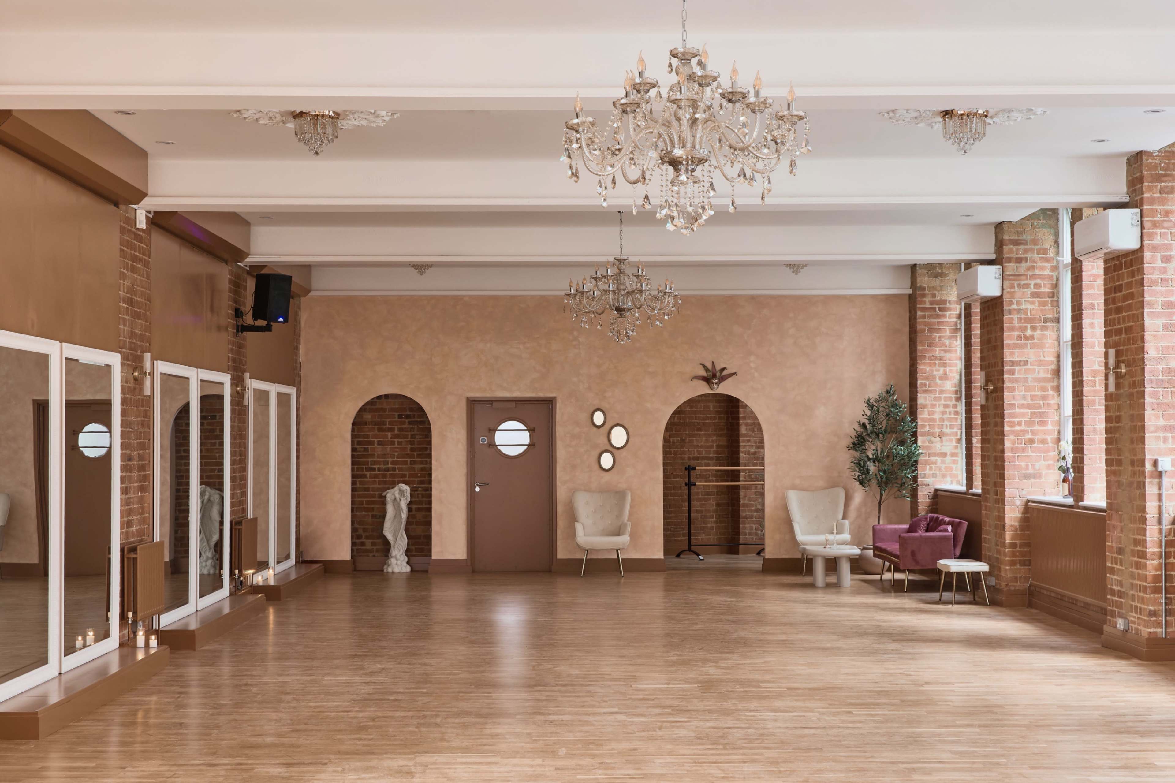 The image shows a spacious, well-lit dance studio featuring wooden flooring, mirrors, chandeliers, and decorative arches.