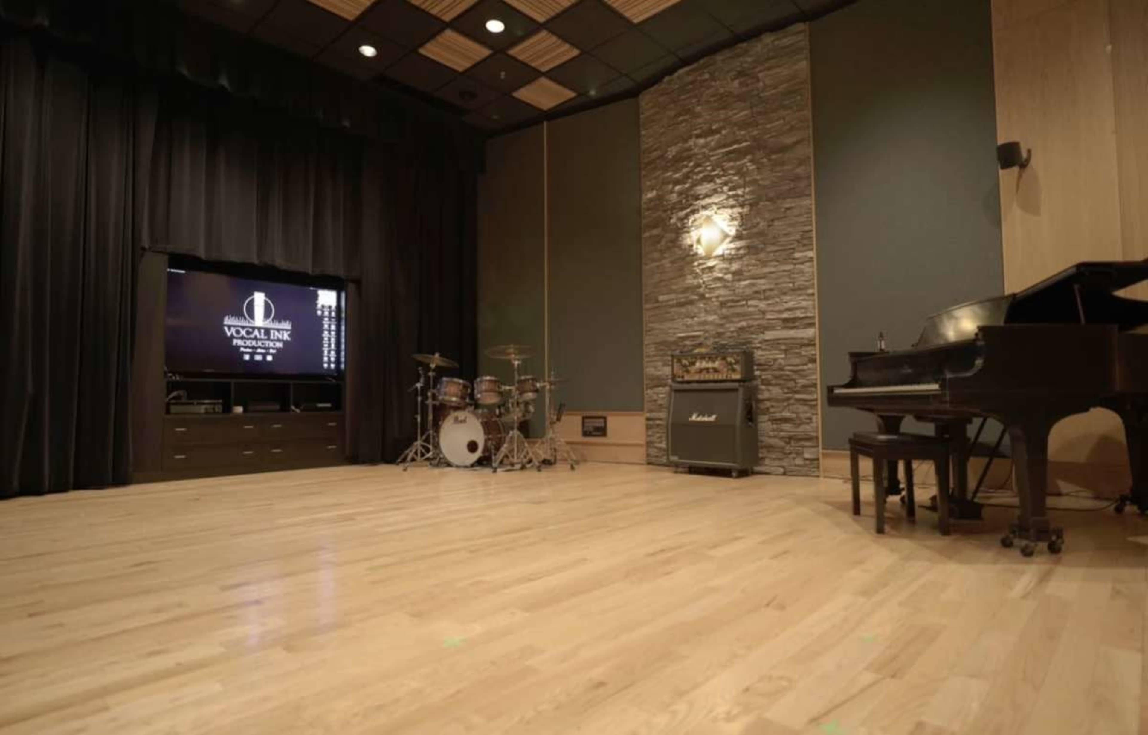 The image shows a music studio with a grand piano, a drum set, and an amplifier against a backdrop of black curtains and a stone wall.