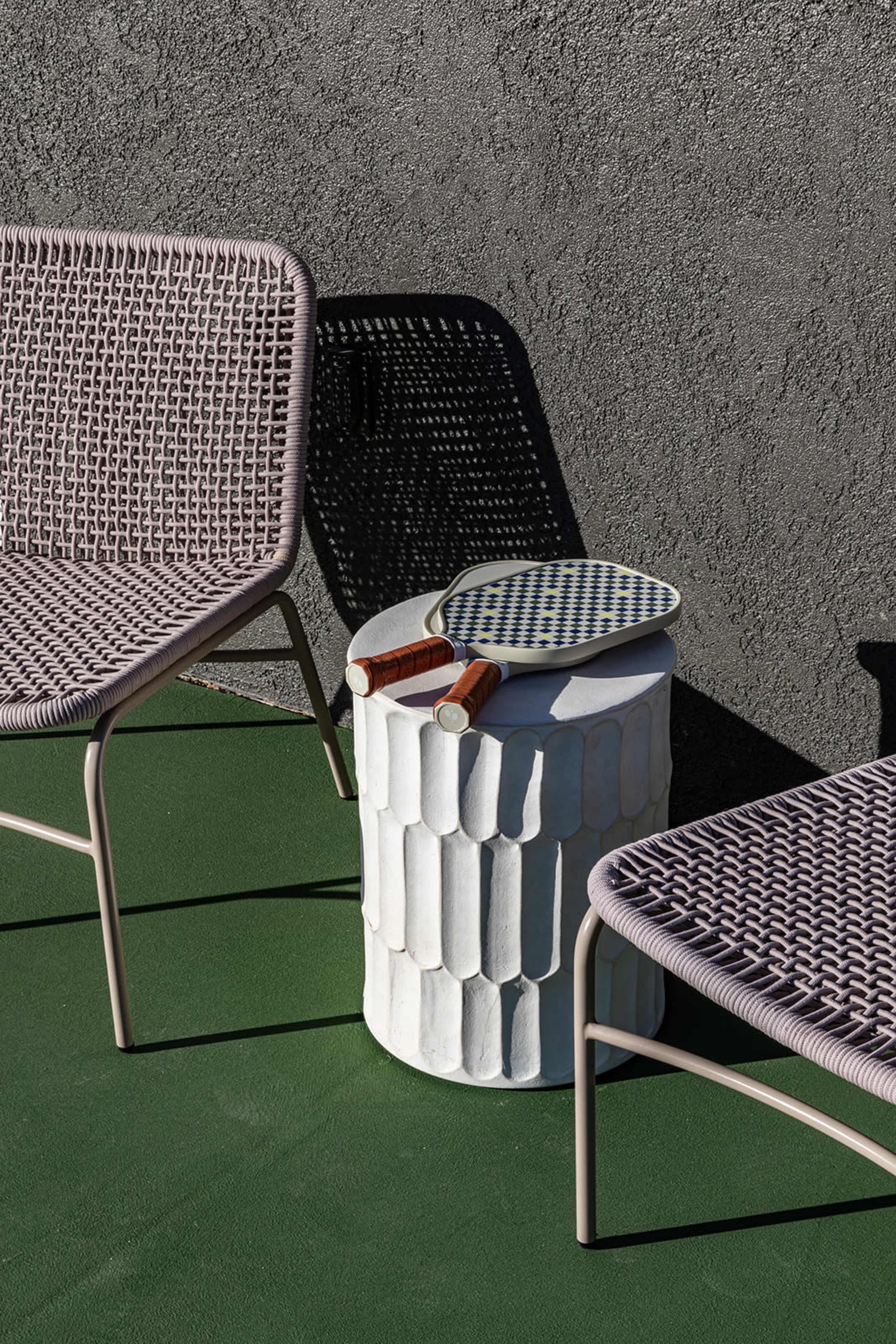 Two woven chairs are positioned beside a textured, white side table topped with a small mirror and rolled items, against a gray wall.