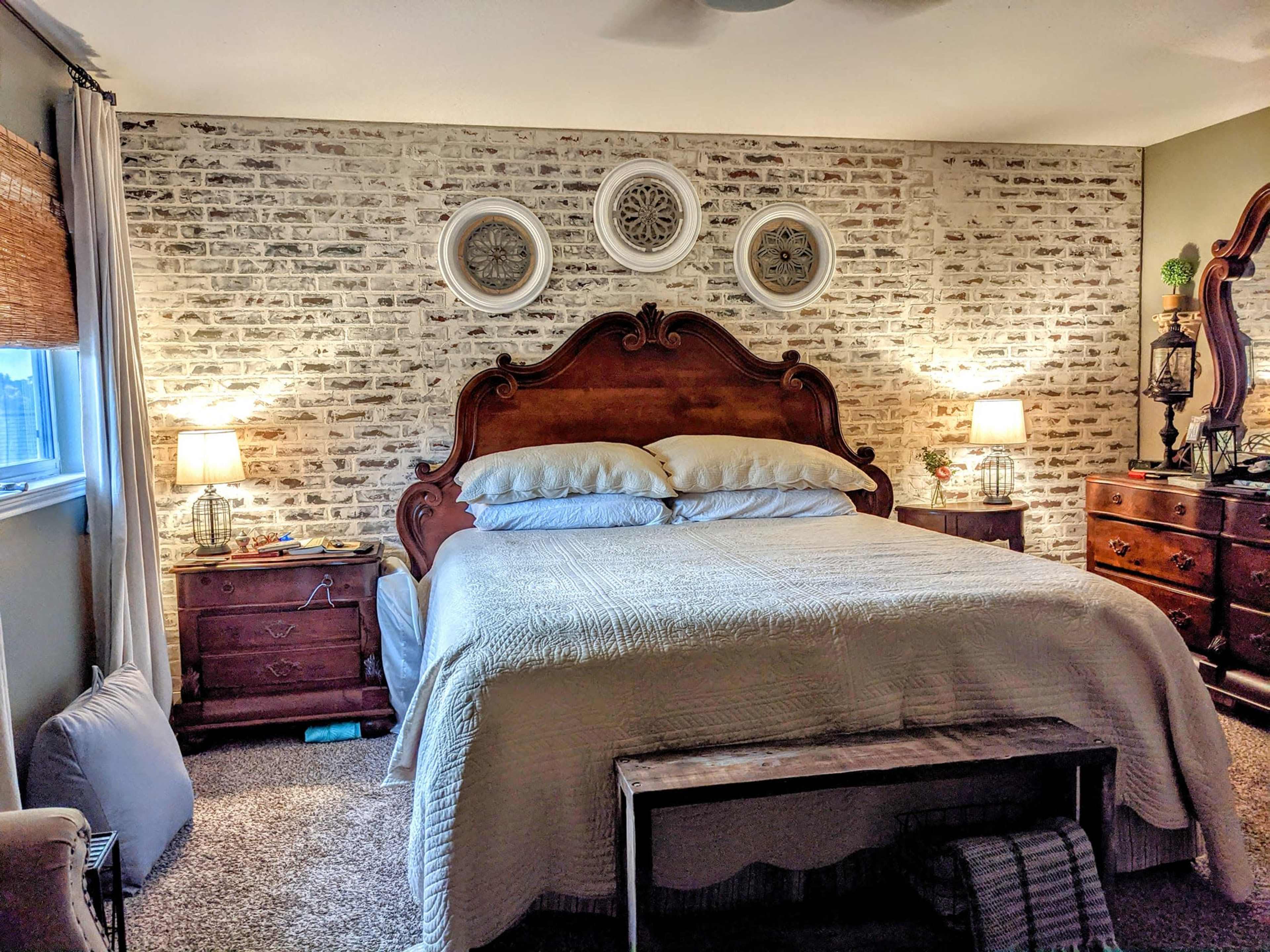 A cozy bedroom features a wooden bed with white bedding, flanked by nightstands and illuminated by lamps, all against a brick-patterned wall.