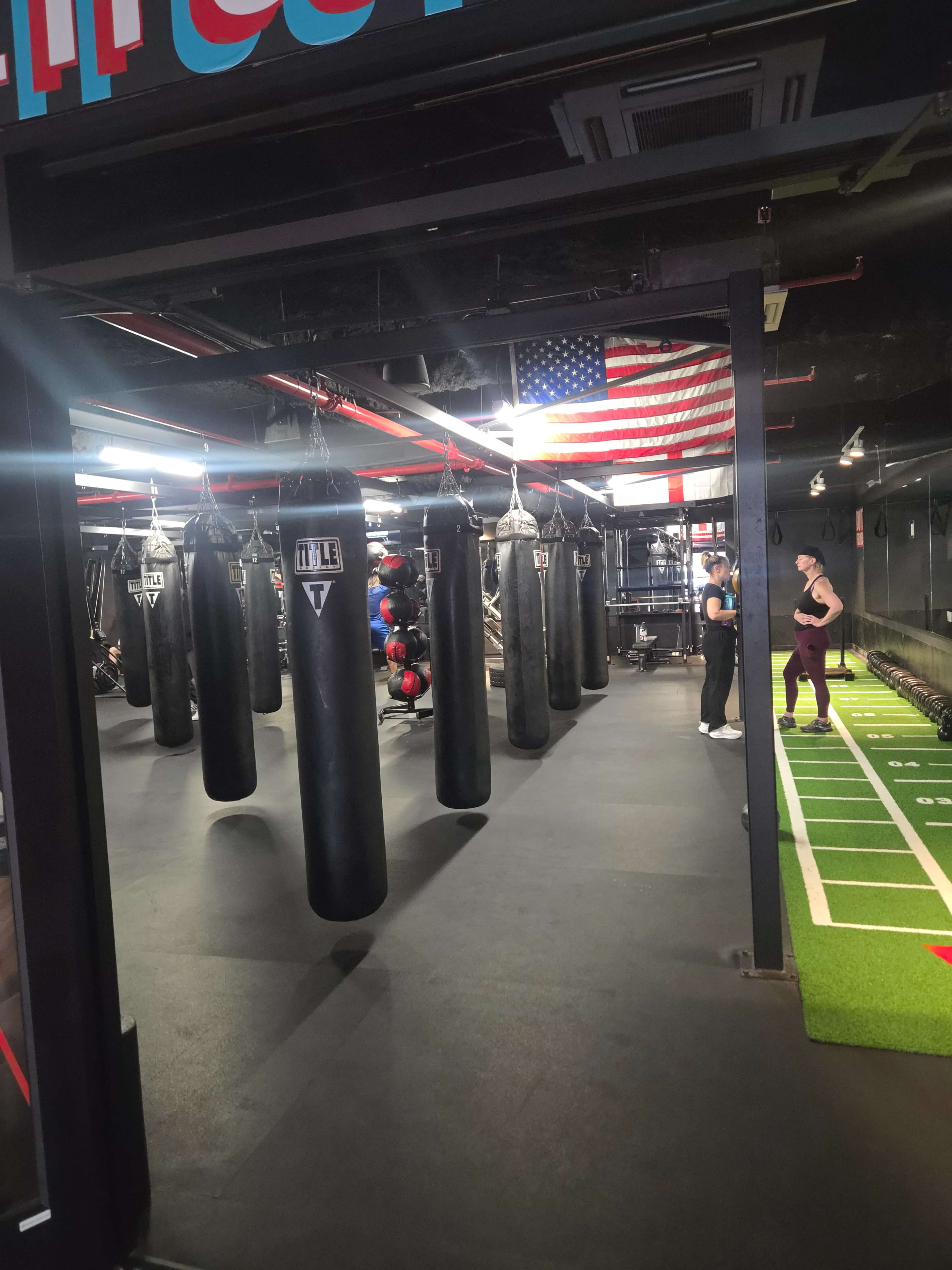 The image shows a fitness gym with several black punching bags hanging from the ceiling, an American flag in the background, and workout equipment along the right side.