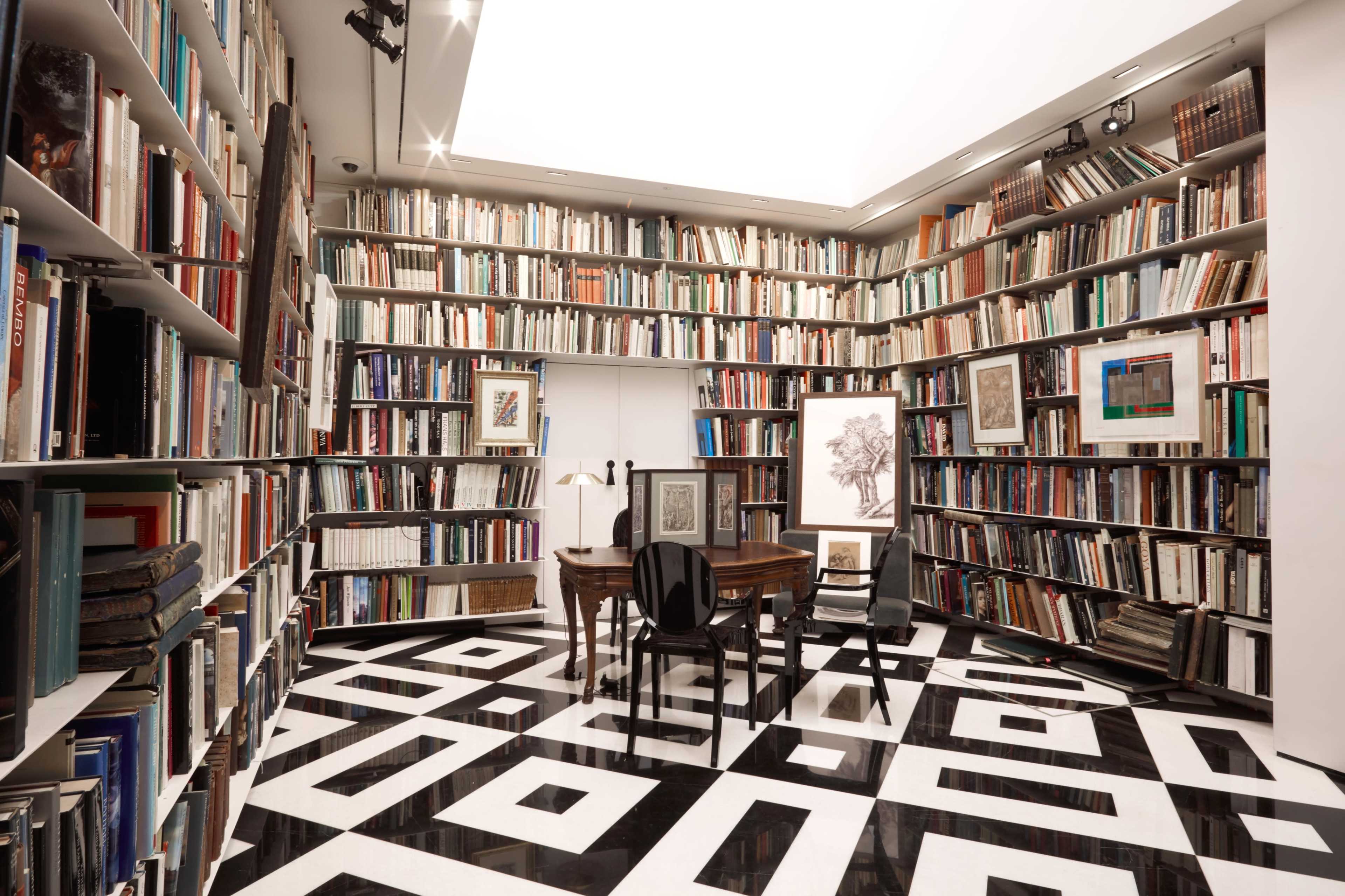 A room filled with tall bookshelves lined with books surrounds a central table and chairs on a patterned black and white floor.