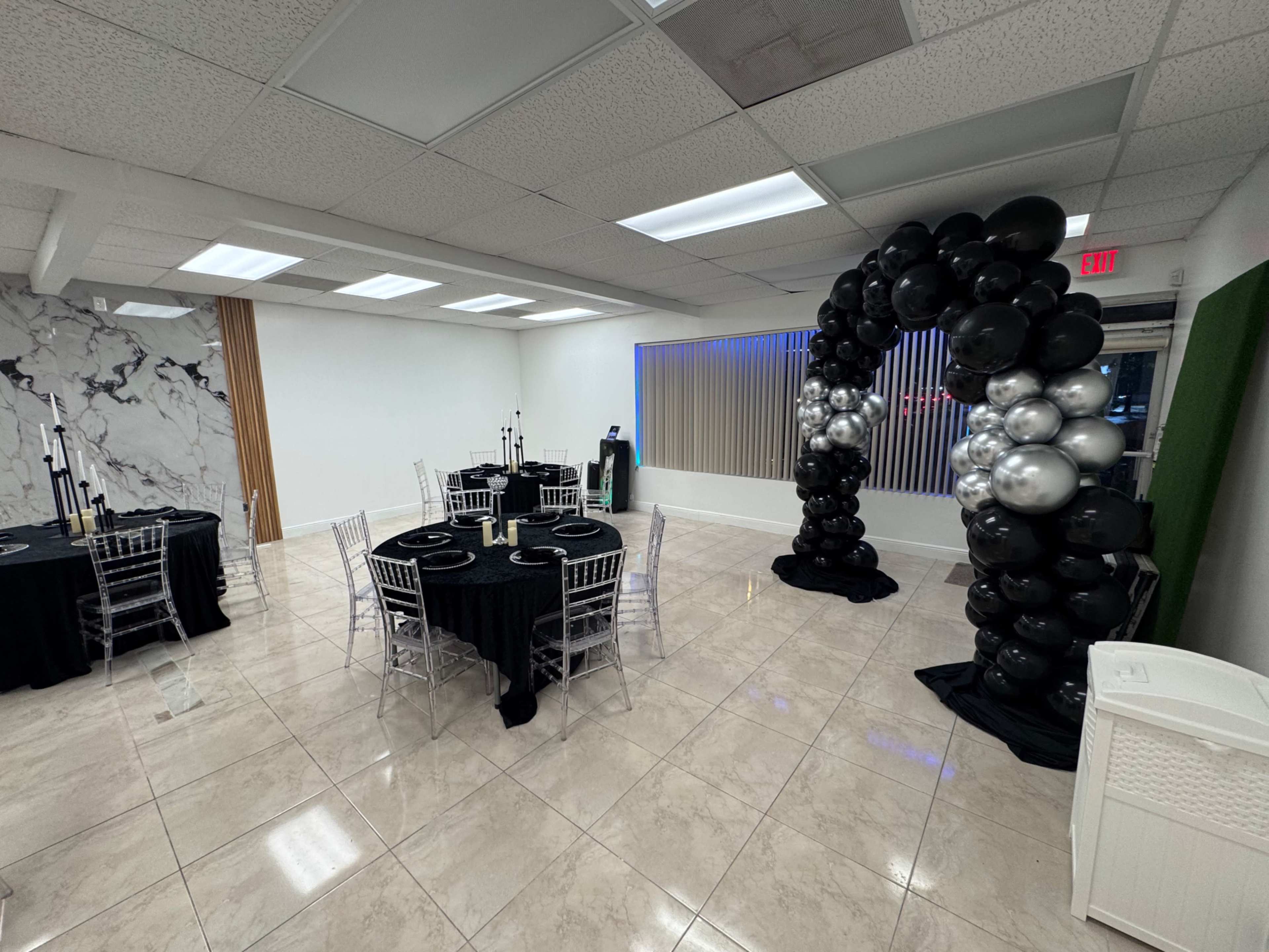 The image shows a spacious event room set up for a gathering, featuring black tables with elegant place settings, a decorative balloon arch, and a green wall accent.