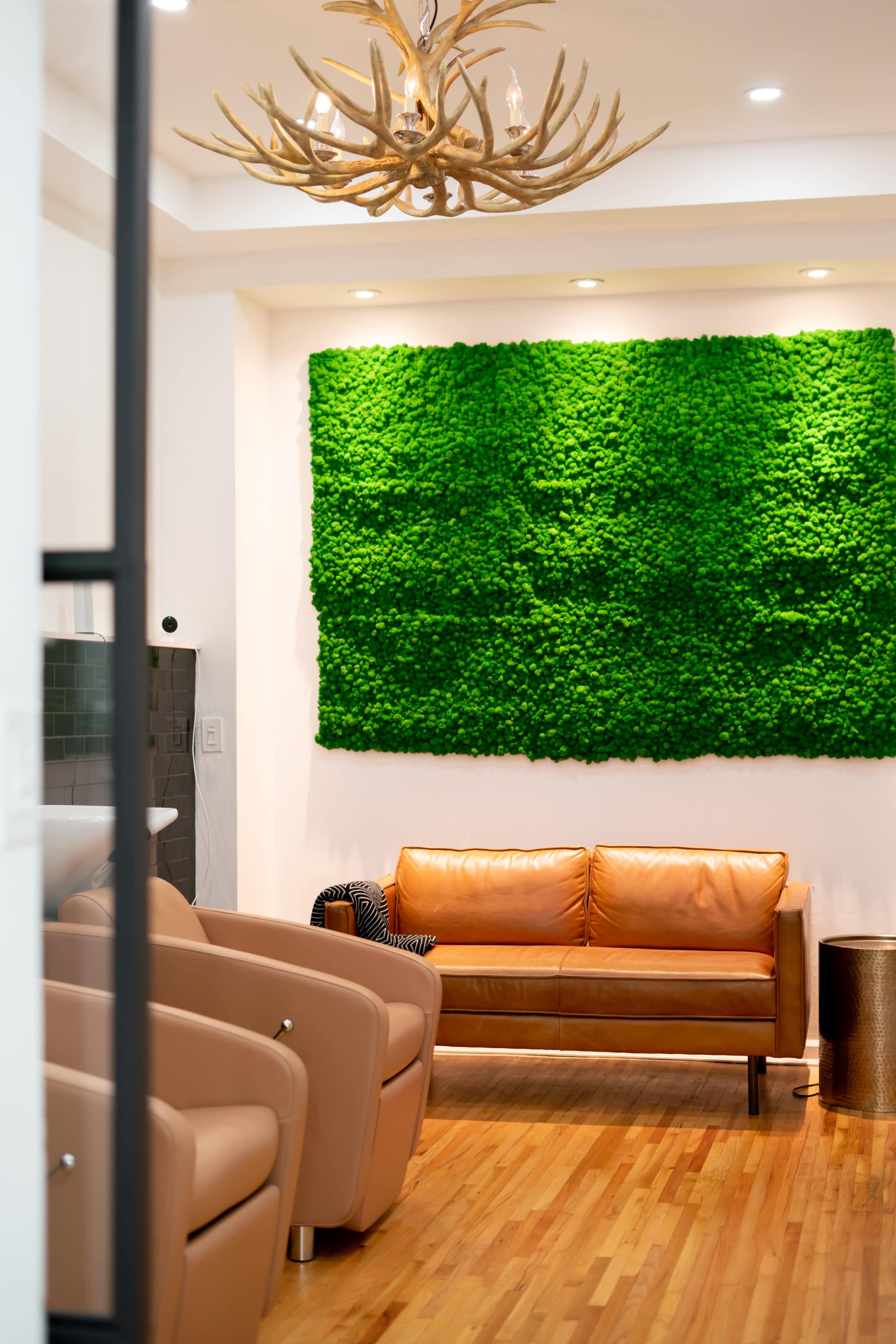 A modern interior with a brown leather couch, beige armchairs, and a large green moss wall mounted behind the couch.