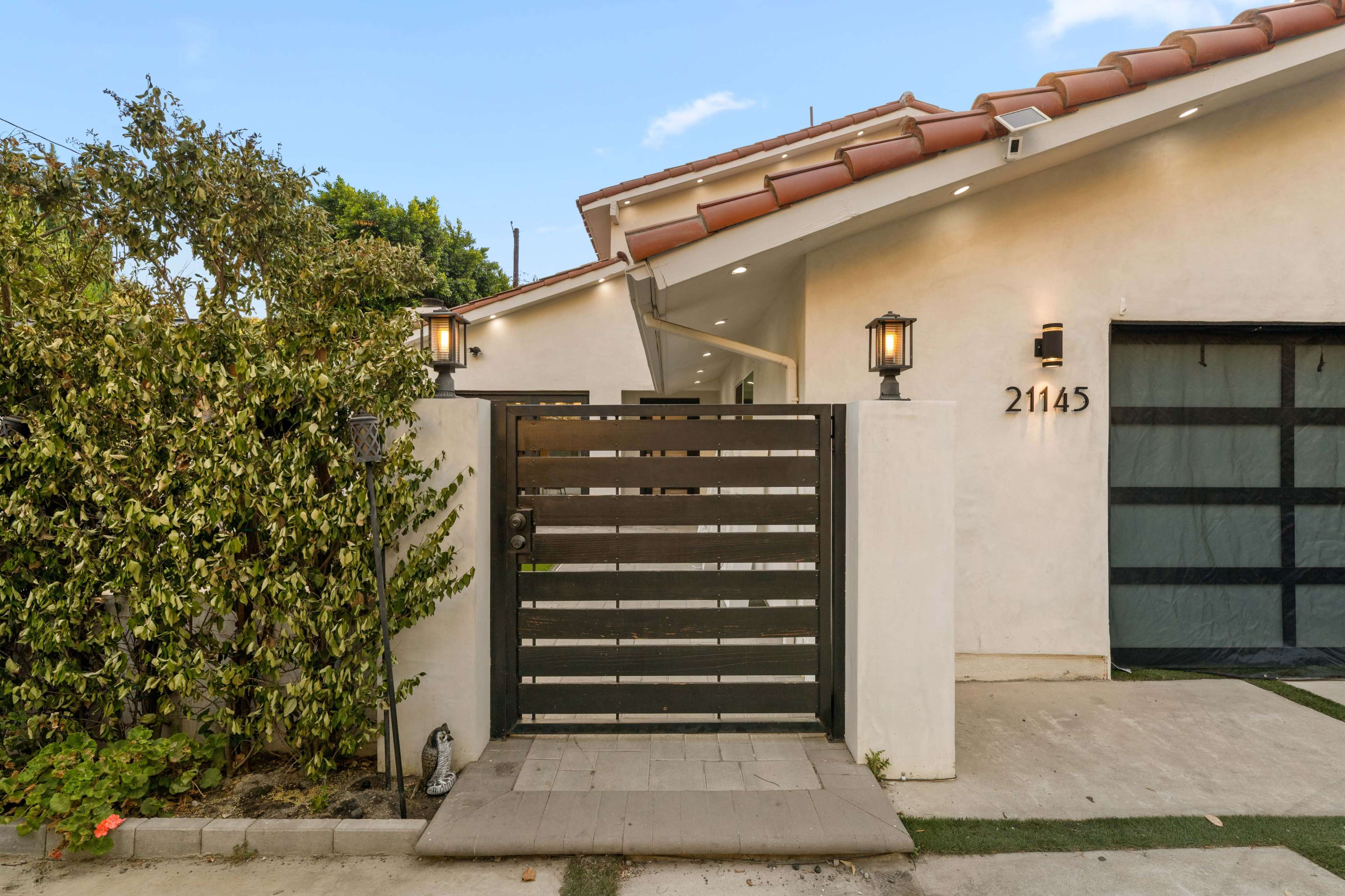 The image shows a modern home entrance with a black horizontal slat gate, a light fixture on either side, and a wall covered in greenery.