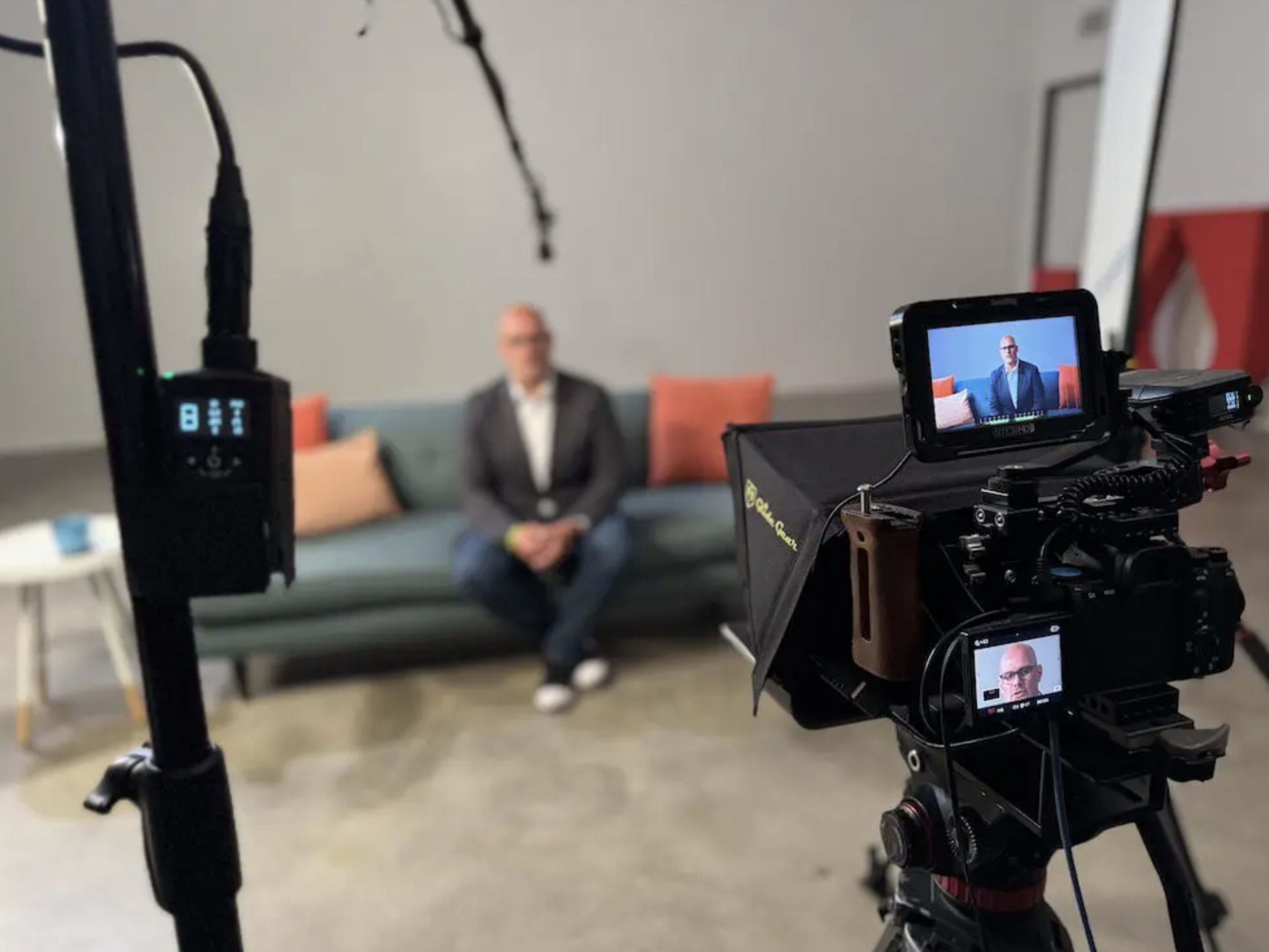 A man in a blazer sits on a couch while two cameras capture the scene in a studio setting.