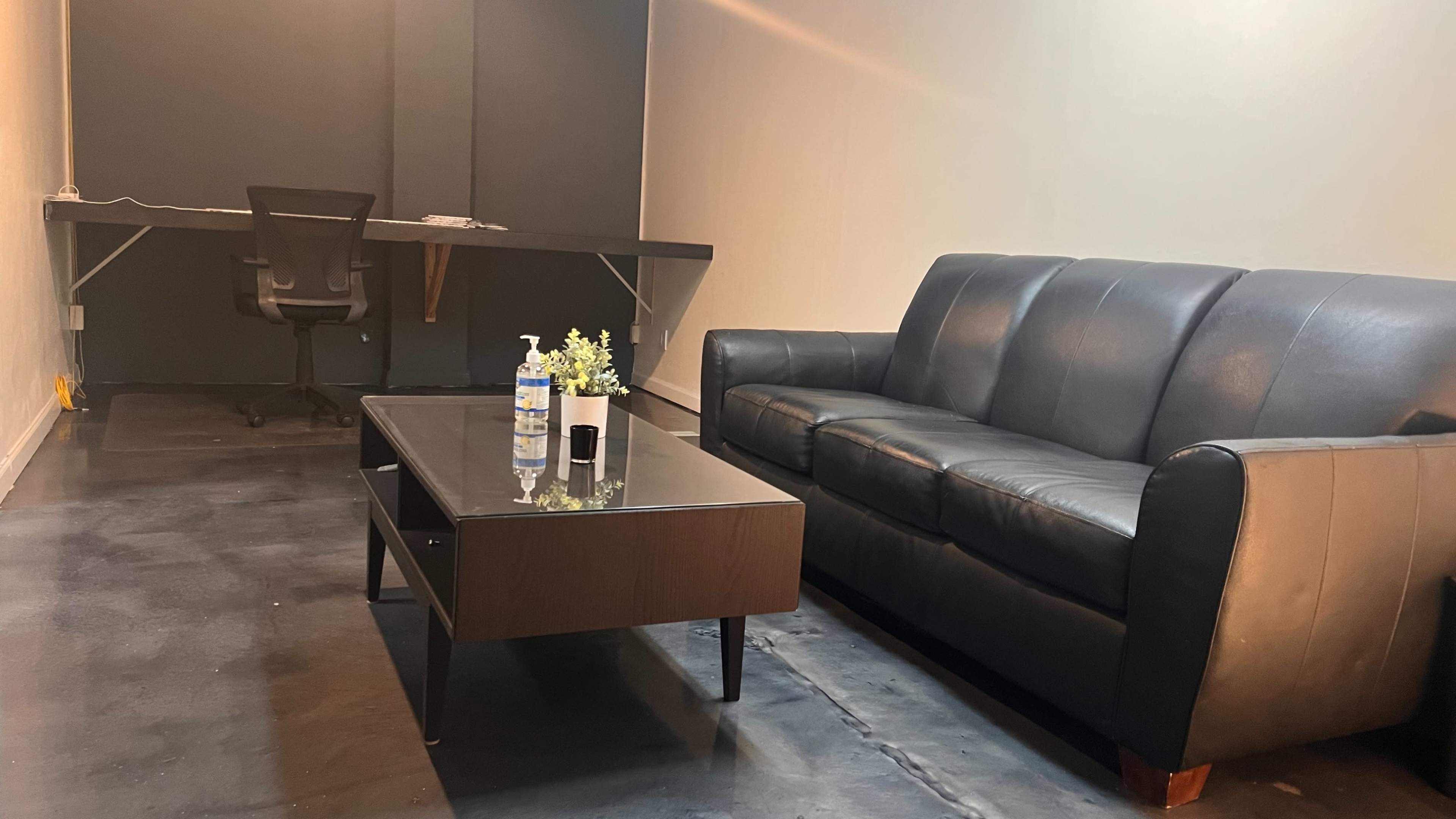 A minimalist room with a black leather couch, a coffee table, and a desk with a chair against a plain wall.