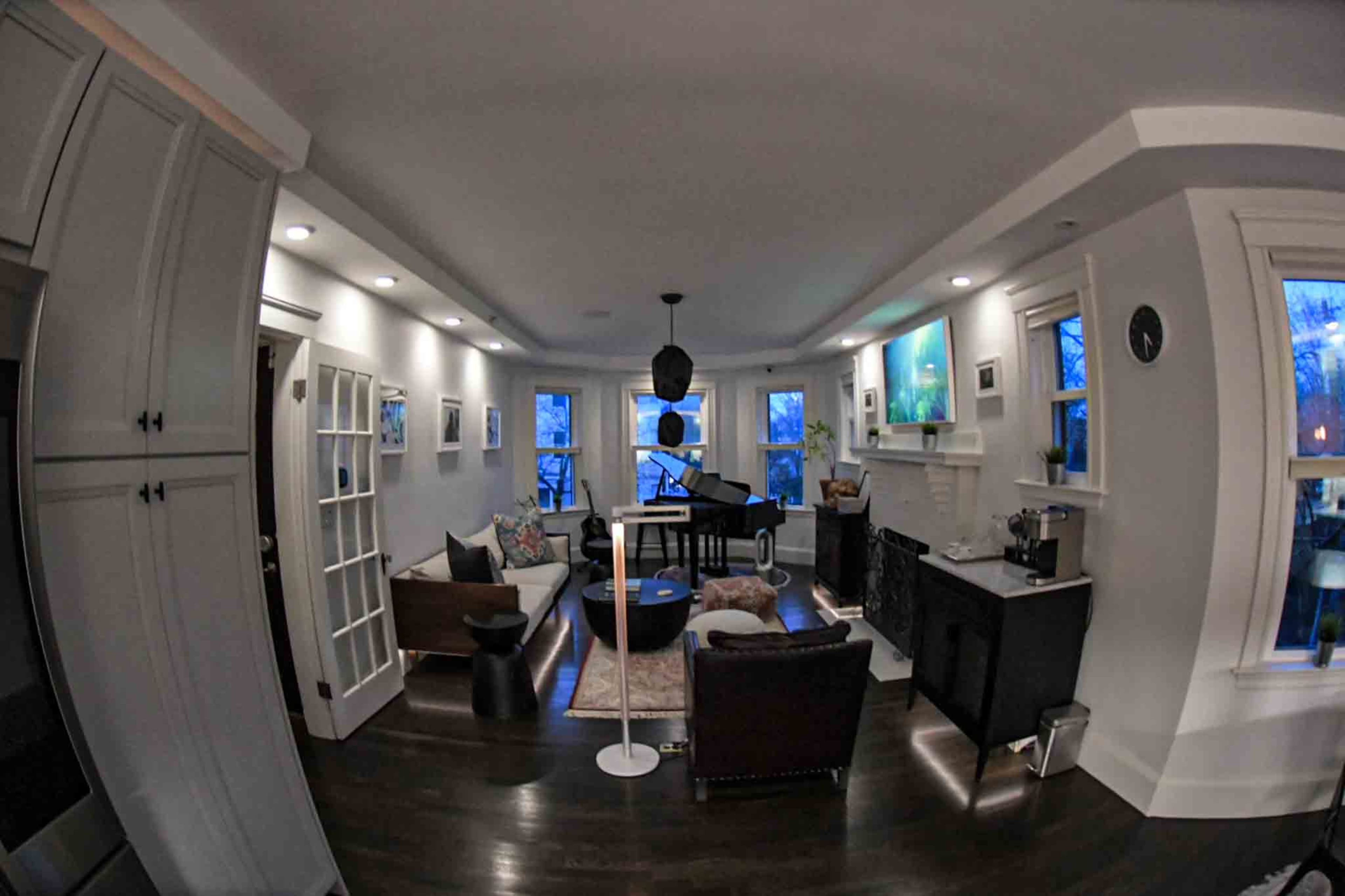 The image shows a modern living room featuring a piano, comfortable seating, and large windows providing natural light.