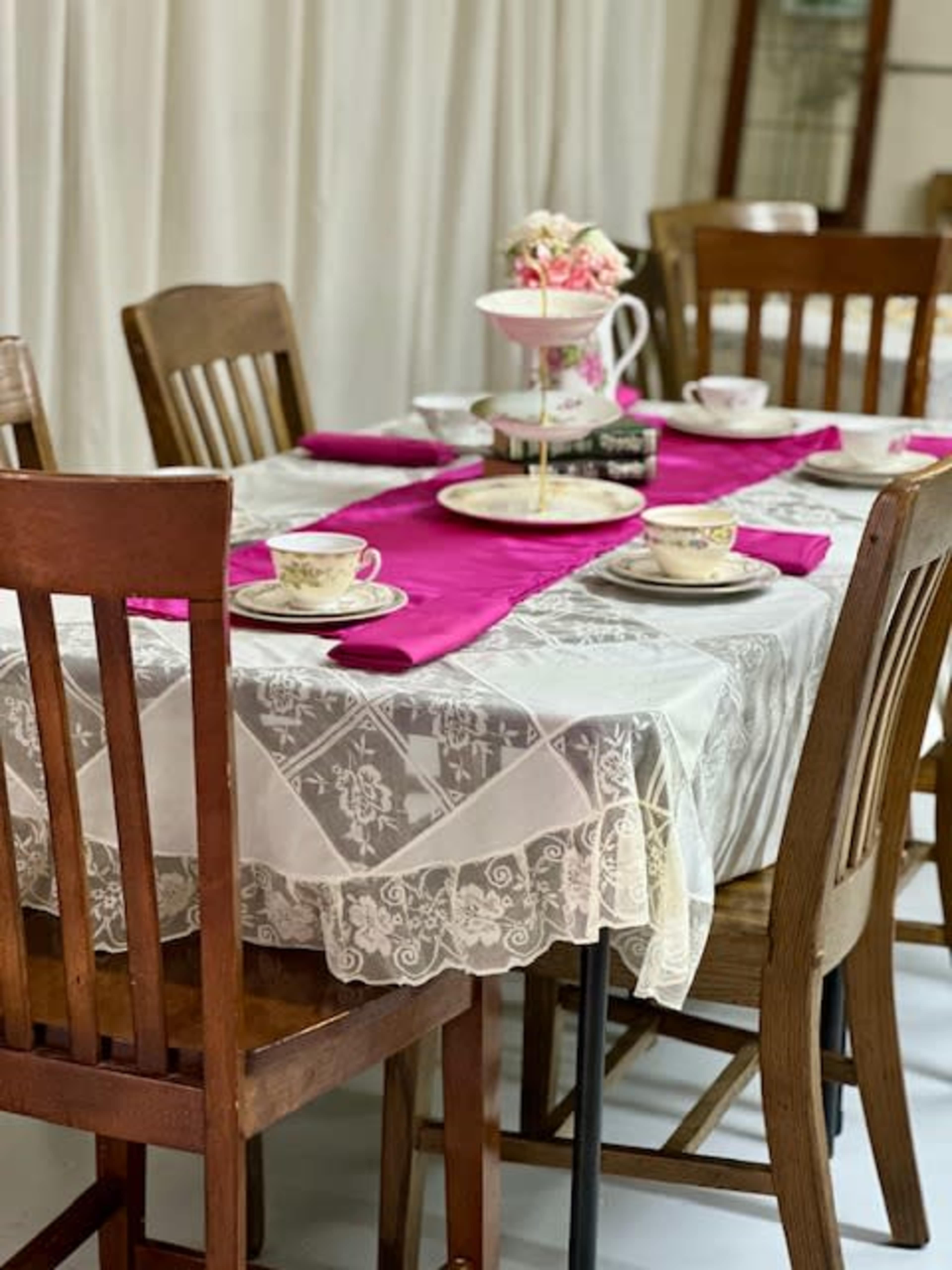 A dining table is set with lace tablecloth, pink placemats, and fine china, surrounded by wooden chairs.