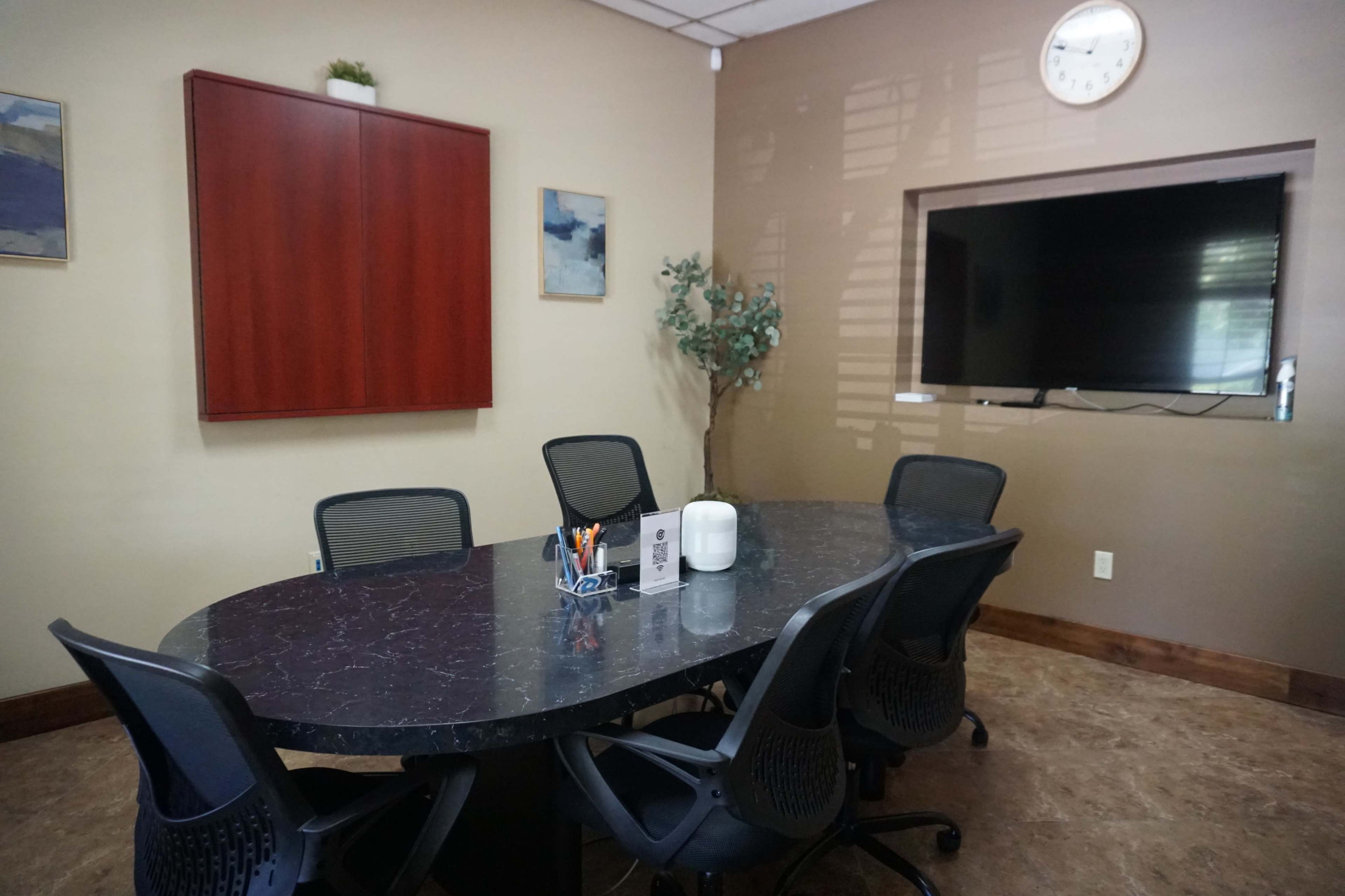 A conference room features a large oval table surrounded by black mesh chairs, with a wall-mounted TV and artwork in the background.
