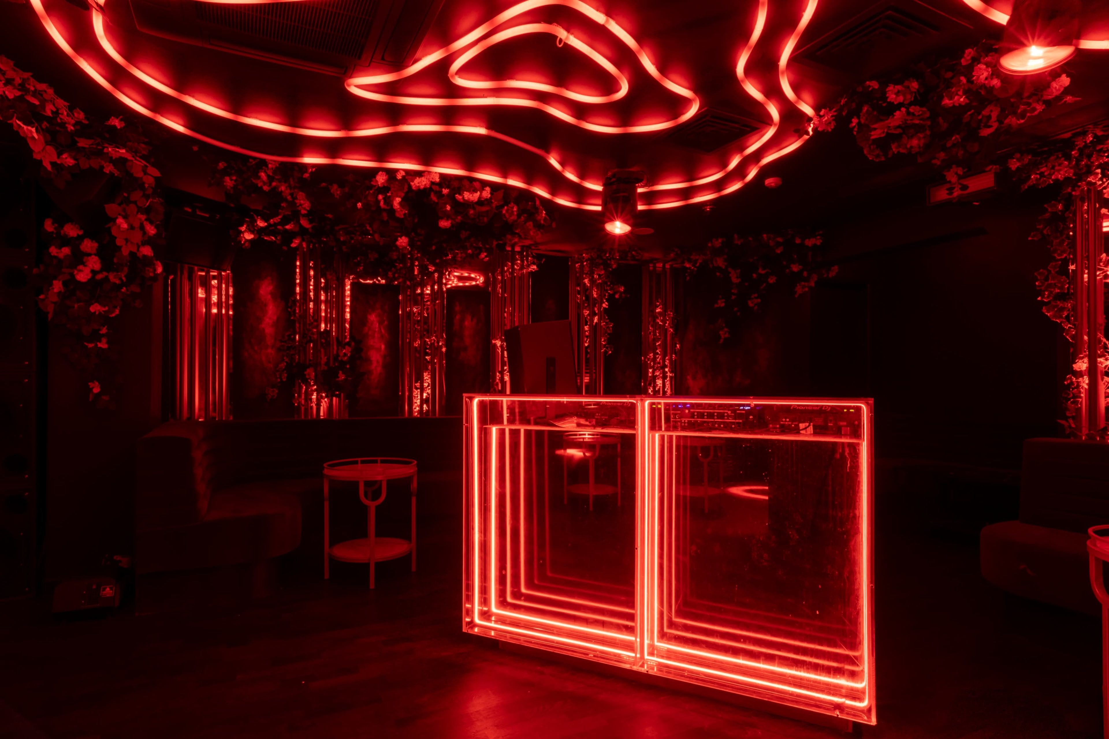 The image shows a dimly lit bar area with red neon lights outlining the bar and decorative elements, surrounded by floral arrangements.