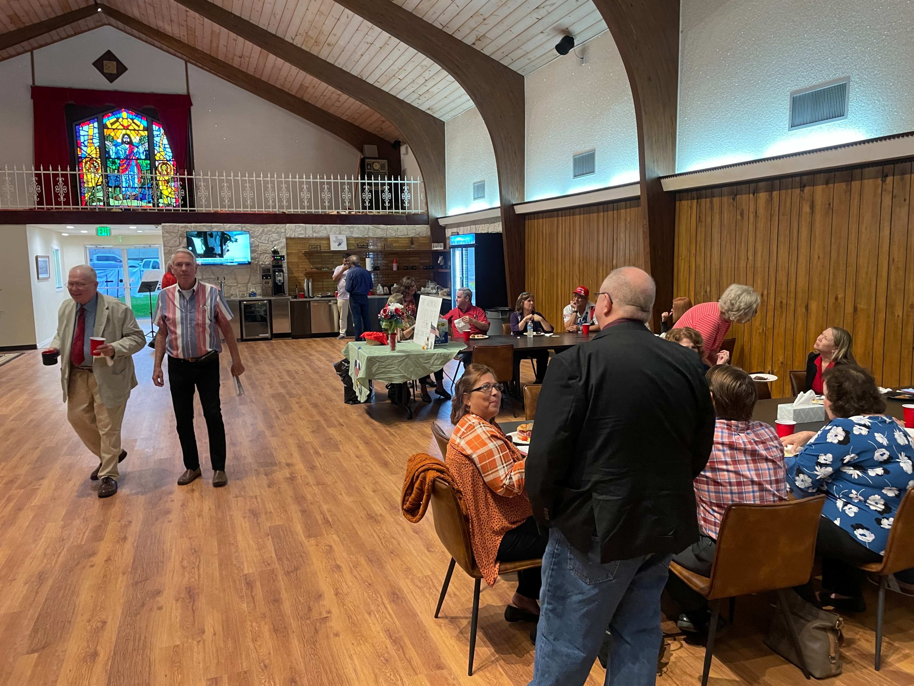 A gathering of people is taking place in a hall, with tables set up for dining and a stained glass window in the background.