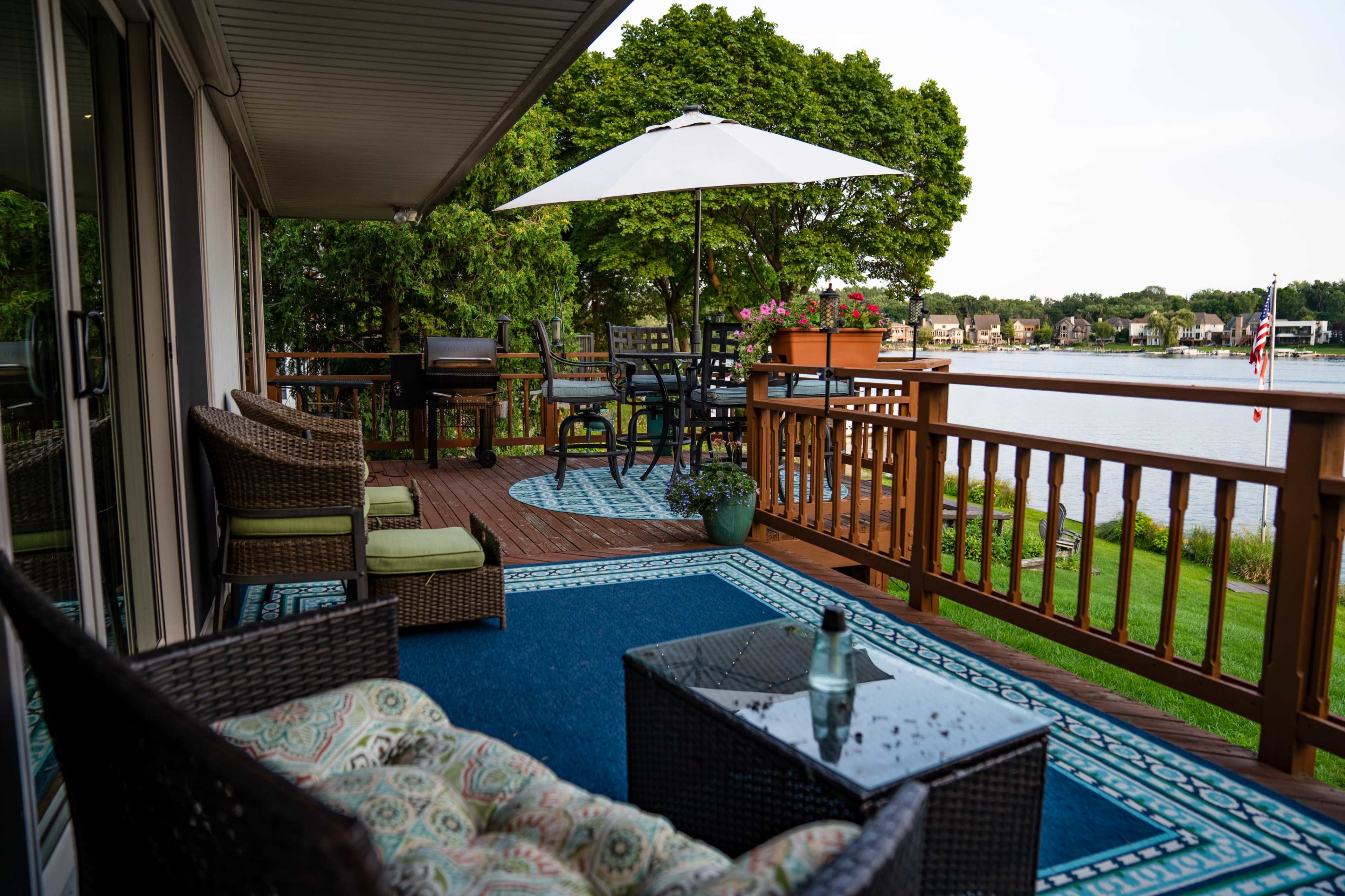 A shaded patio with wicker furniture, a table, and potted plants overlooks a calm lake.