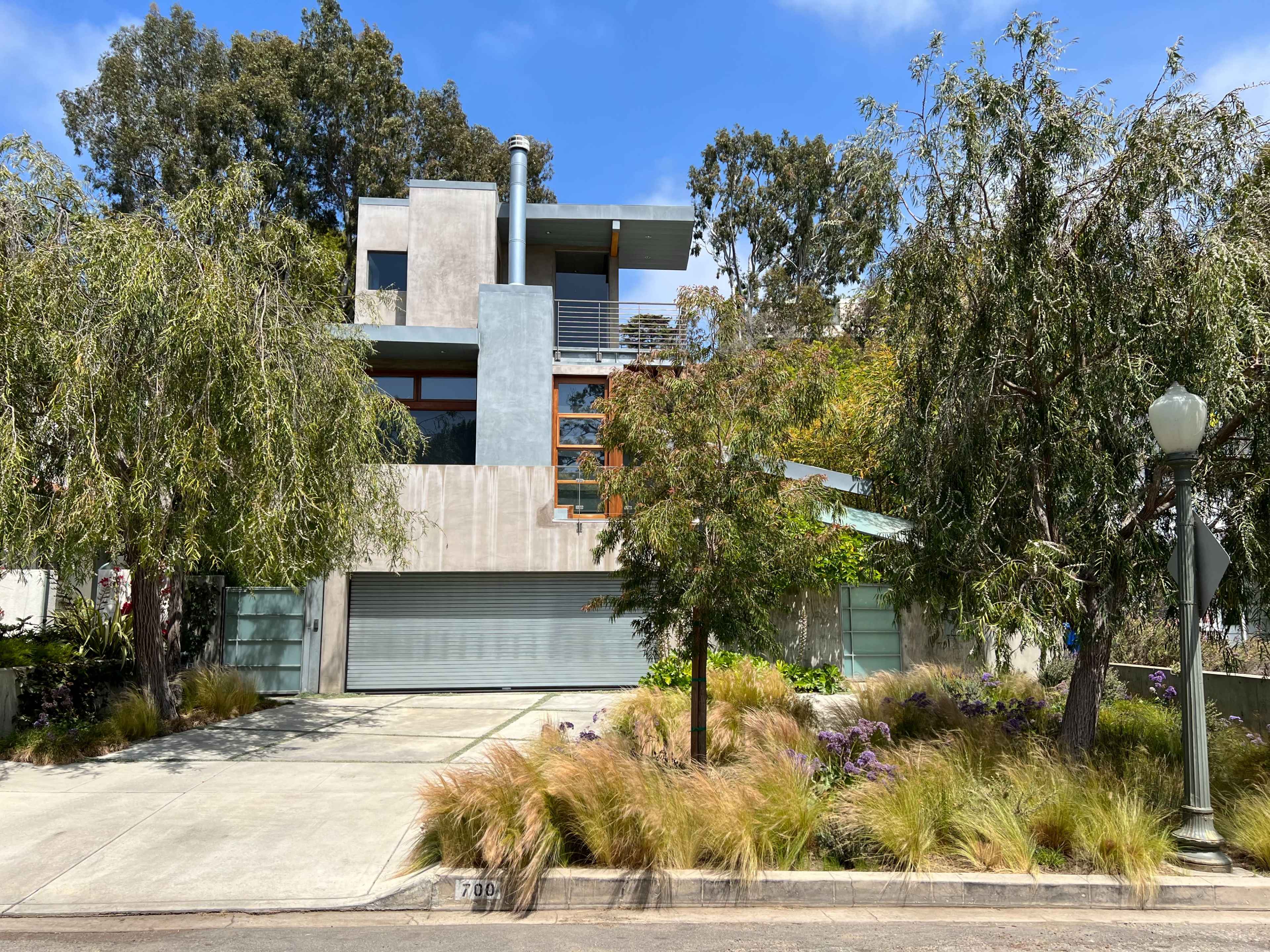 A modern house with a mix of concrete and wood features is set back from the street, surrounded by decorative landscaping and tall trees.