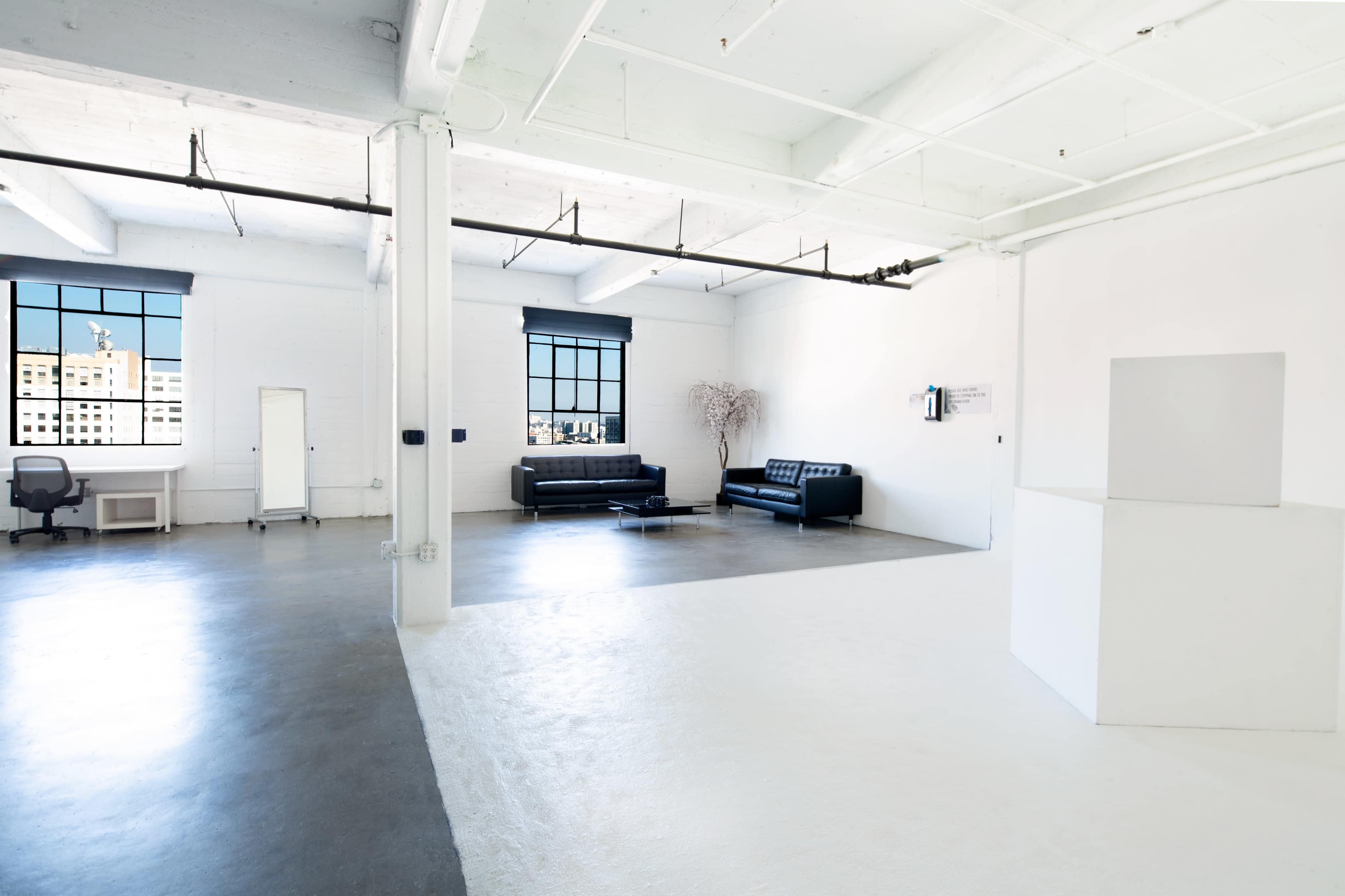 The image depicts a spacious, minimalist room featuring large windows, a black sofa, a desk with a chair, and a white pedestal on a divided floor.