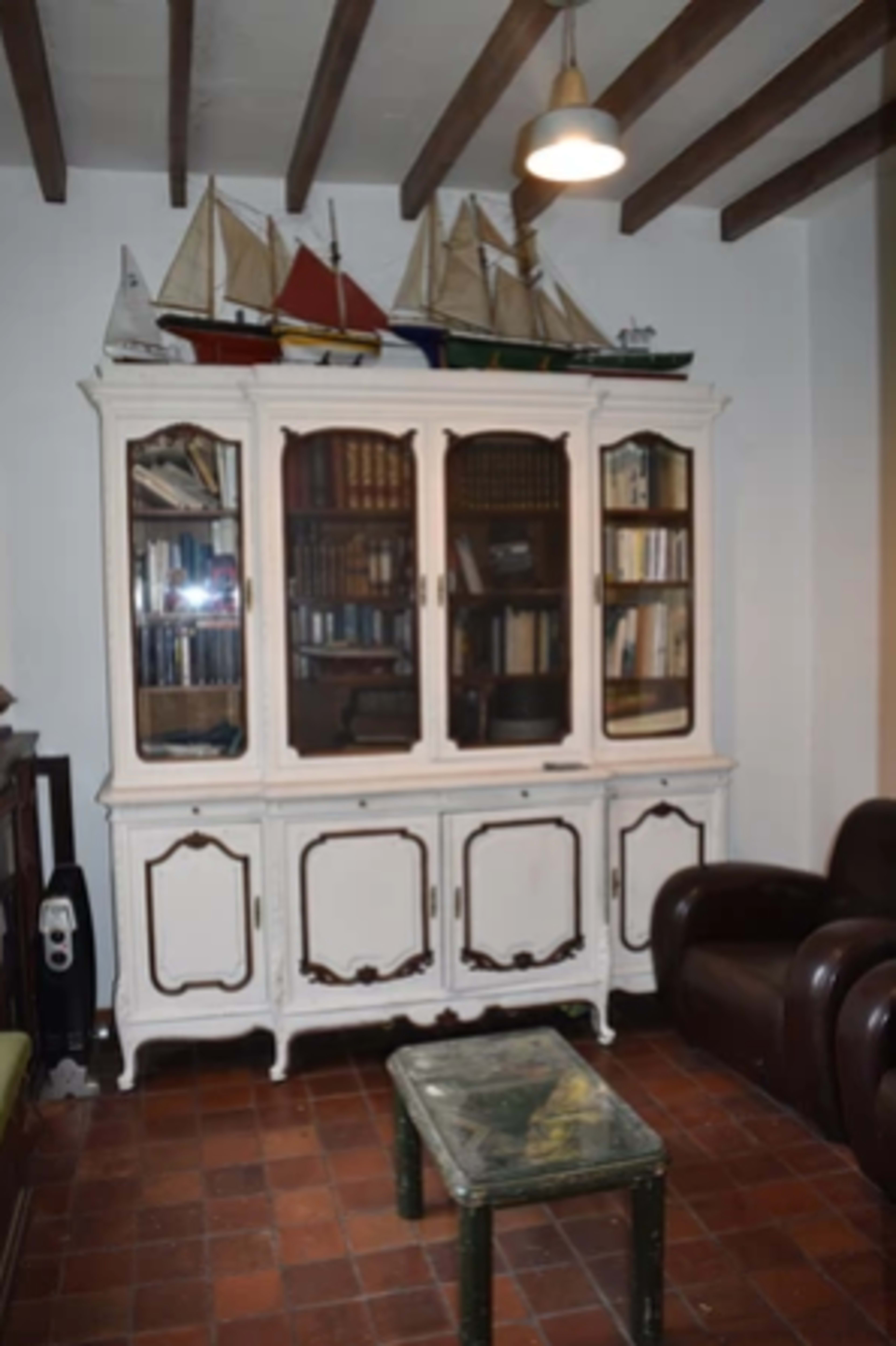 A white wooden cabinet with glass doors displaying books and decorative model sailboats on top, set in a room with a brown chair and a small green table.