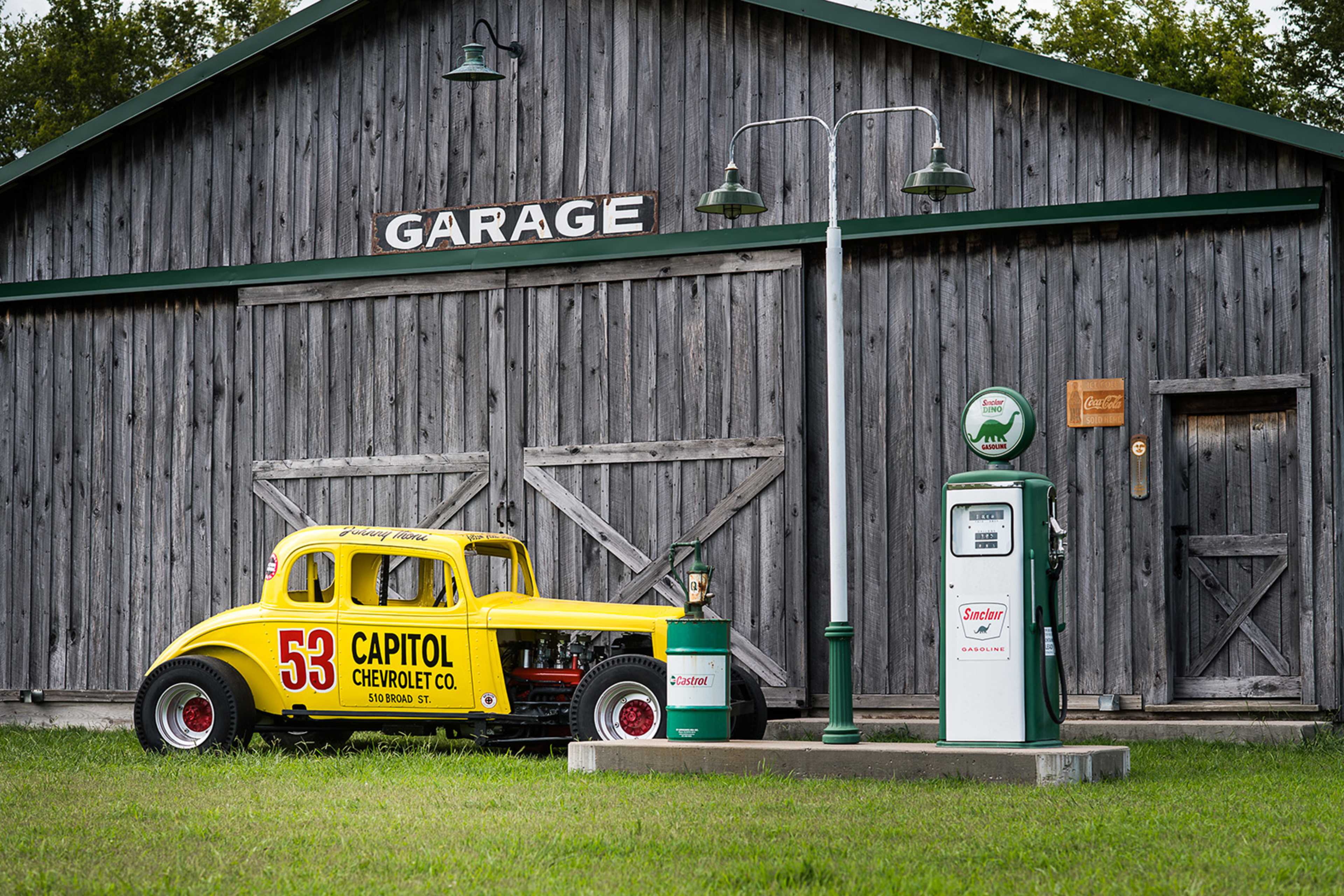 A yellow vintage race car with the number 53 is parked beside a green and white gas pump in front of a rustic wooden garage.