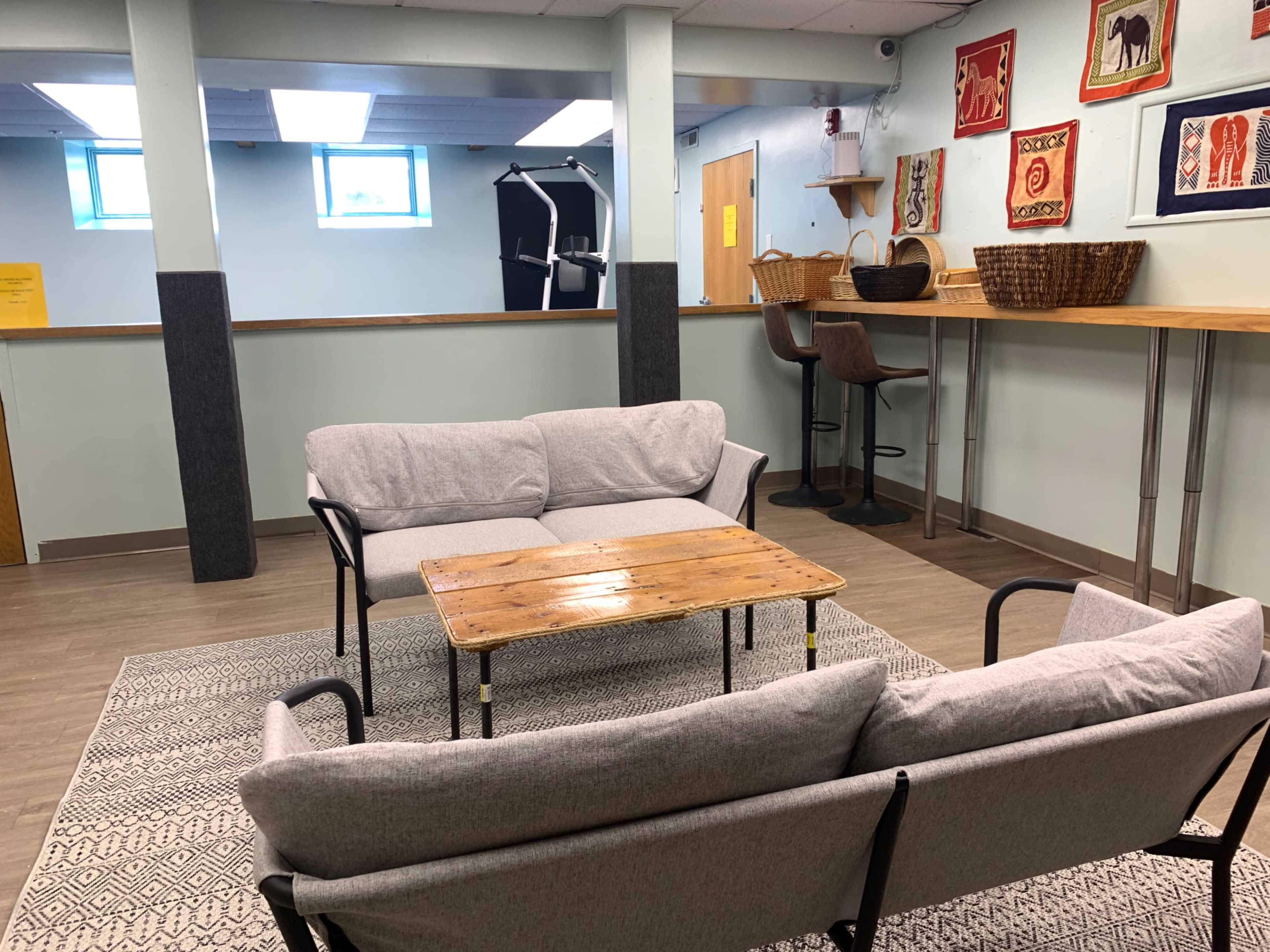The image shows a cozy seating area with two gray sofas and a wooden coffee table, set in a room with light blue walls and a counter displaying baskets and wall decorations.