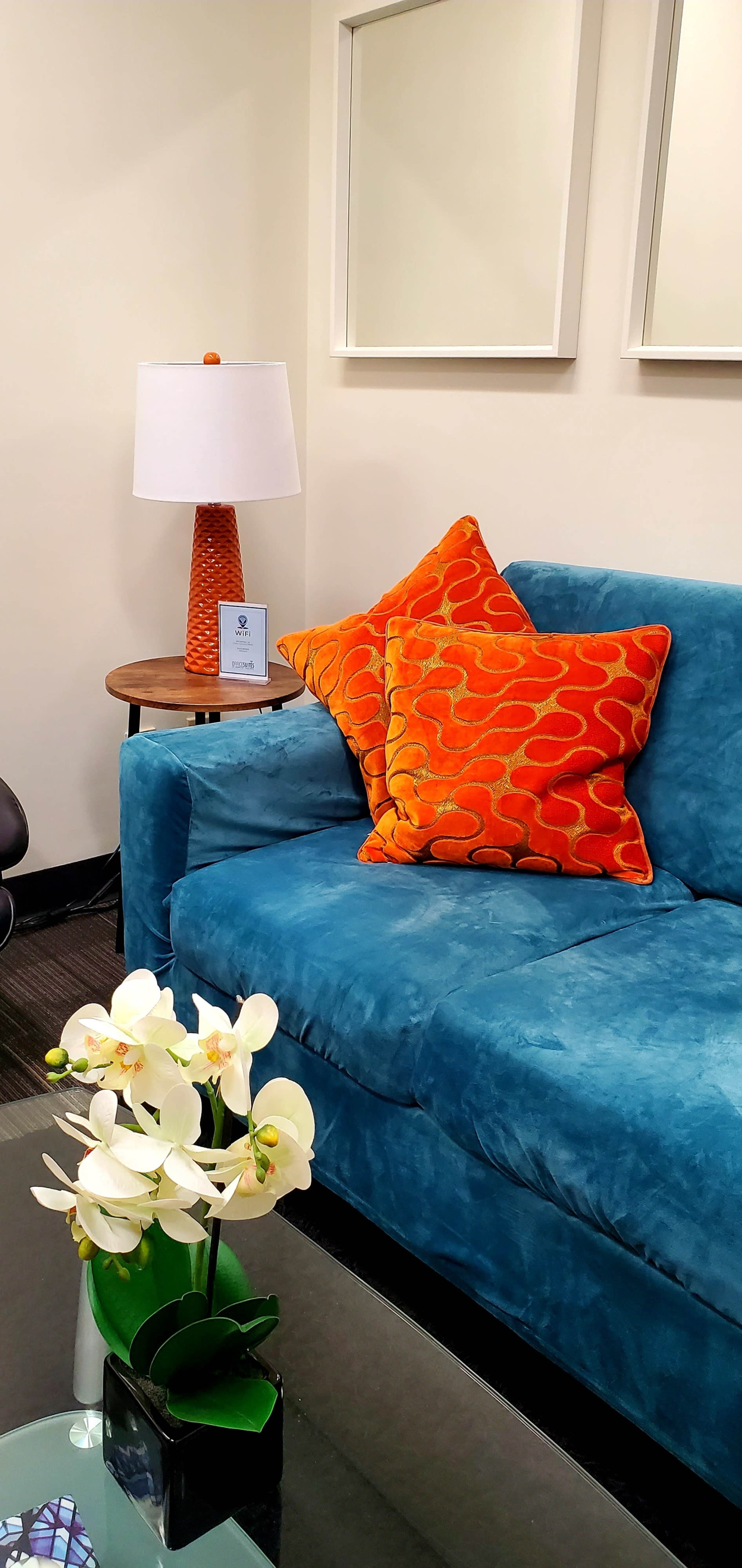 A blue sofa with orange patterned pillows sits next to a lamp and a small table, while a floral arrangement is displayed on the coffee table.