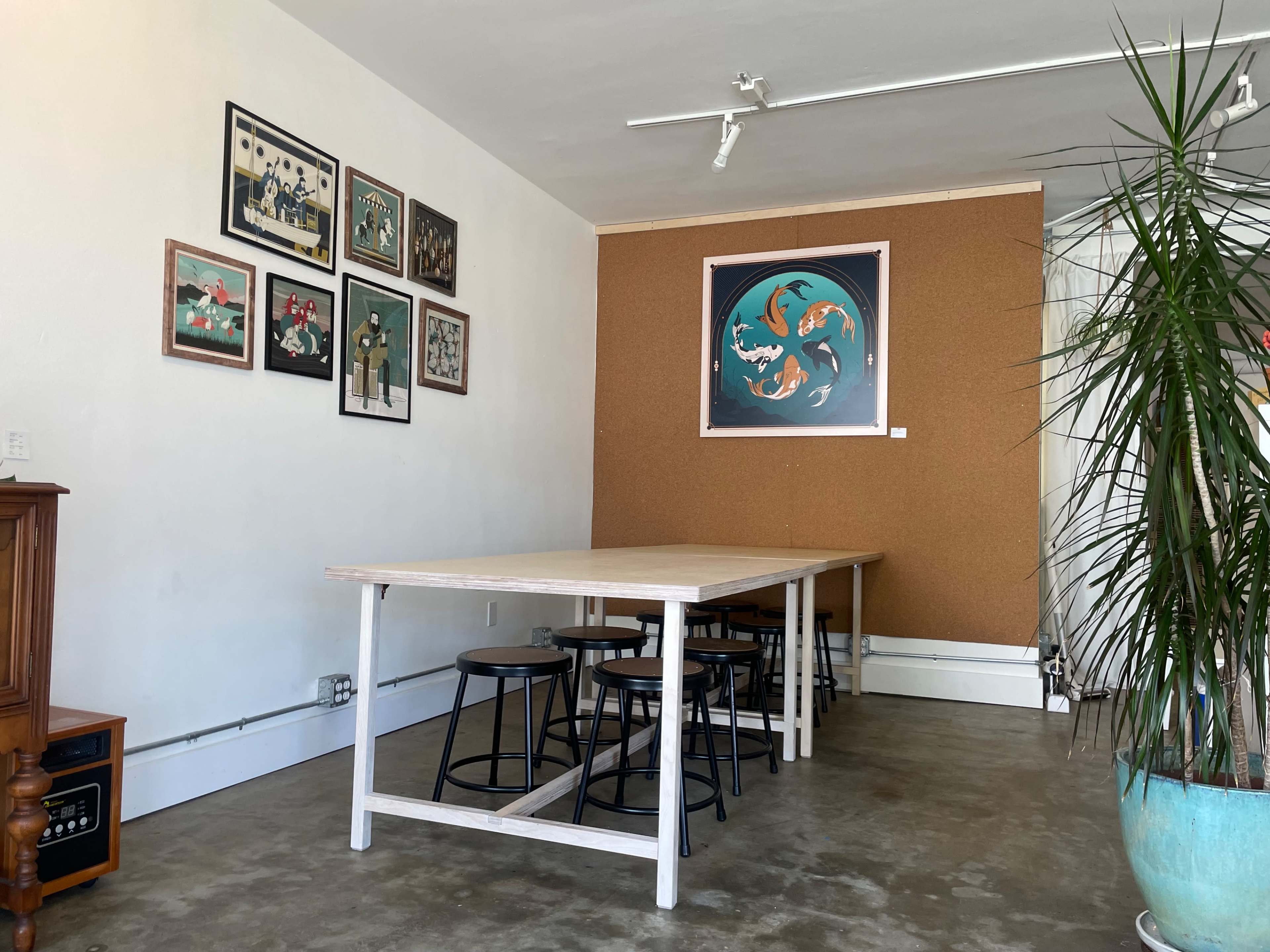 The image shows a minimalist room with a large wooden table surrounded by black stools, alongside a wall featuring framed artworks and a corkboard.