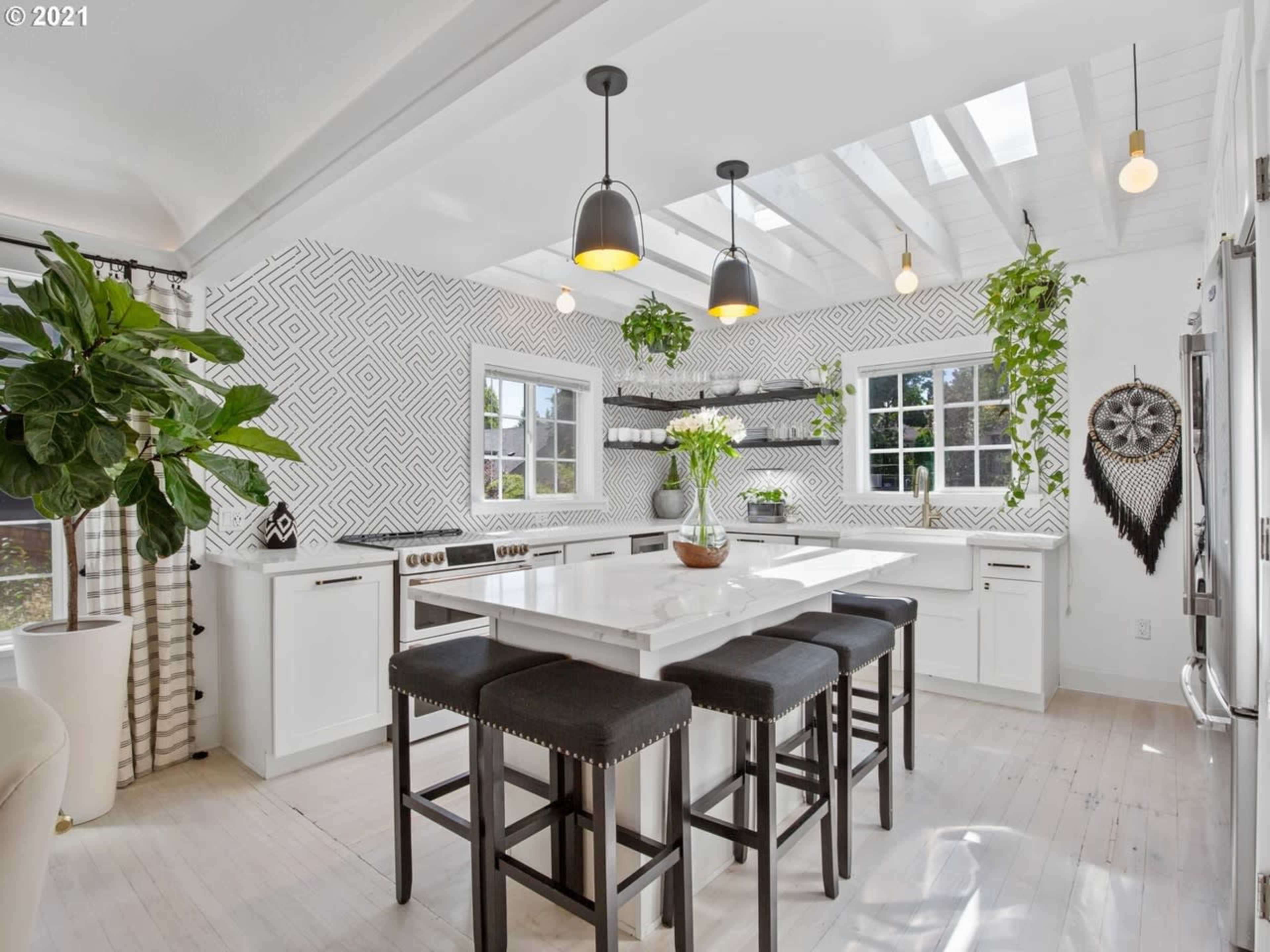 A bright kitchen features a white island with black stools, surrounded by plants and modern lighting, against a geometric-patterned backsplash.
