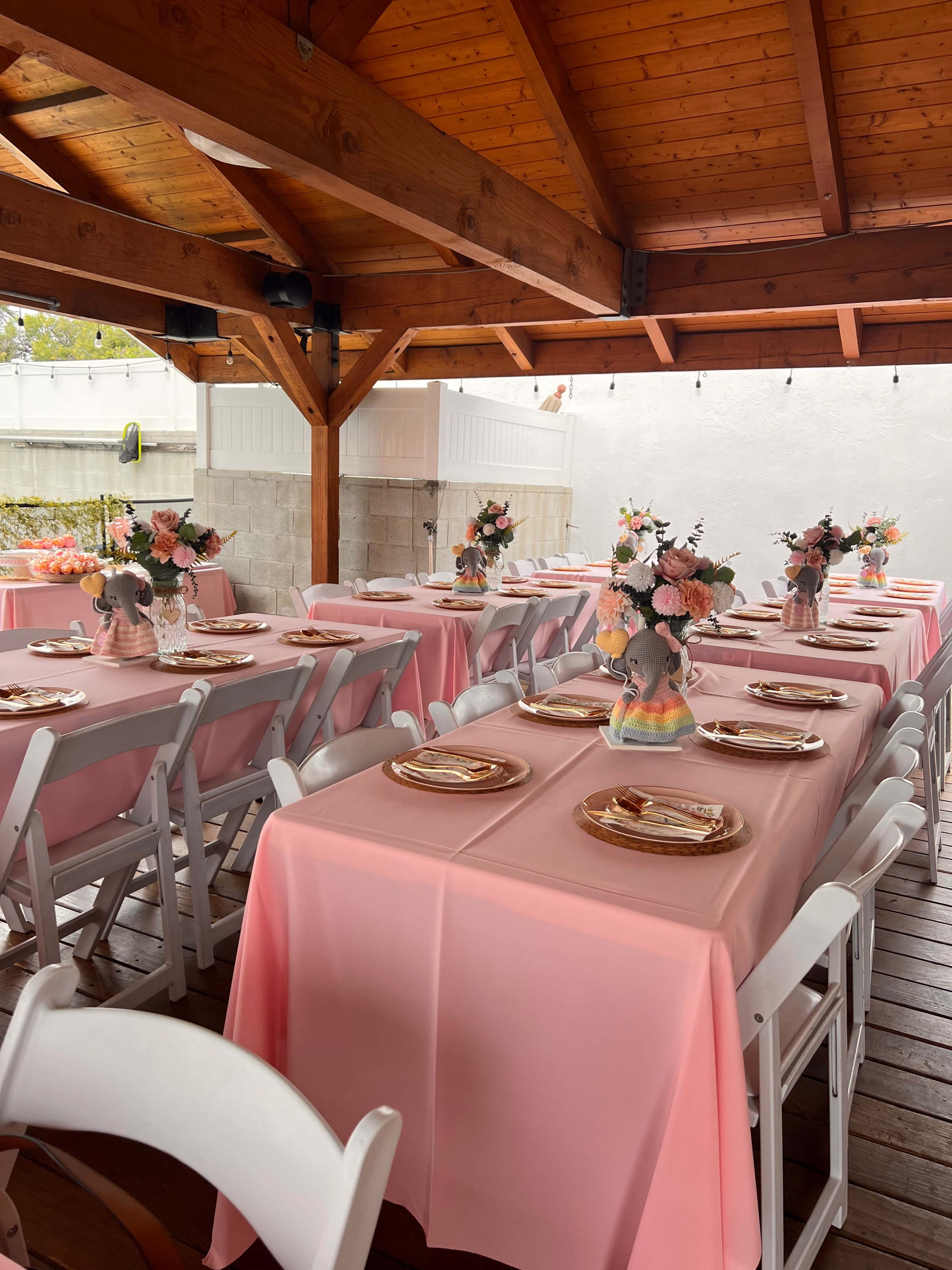The scene shows a decorated outdoor dining area with tables covered in pink tablecloths, arranged with floral centerpieces and place settings.