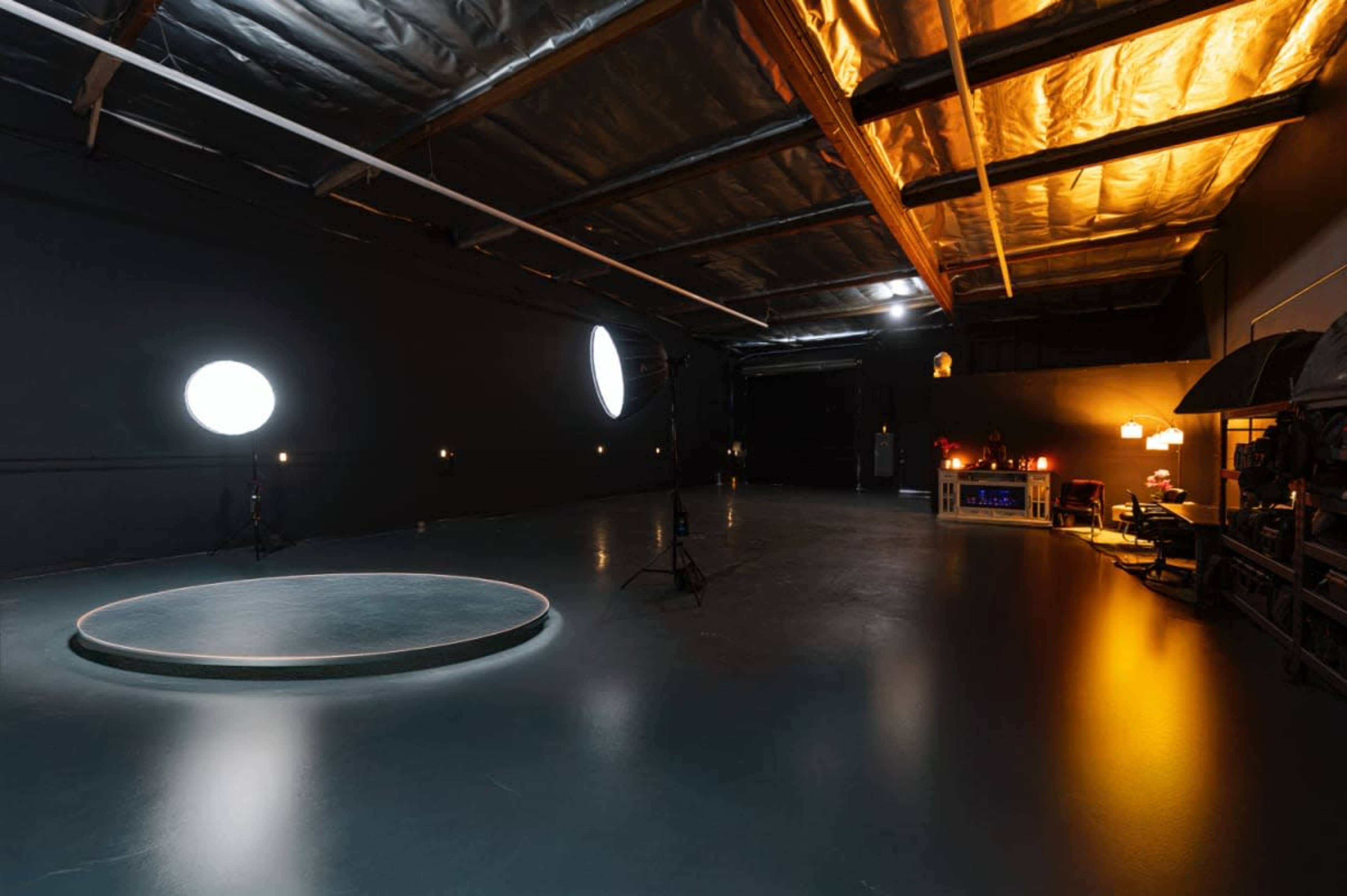 The image shows a spacious studio with a circular platform in the center, surrounded by black walls and various lighting equipment.