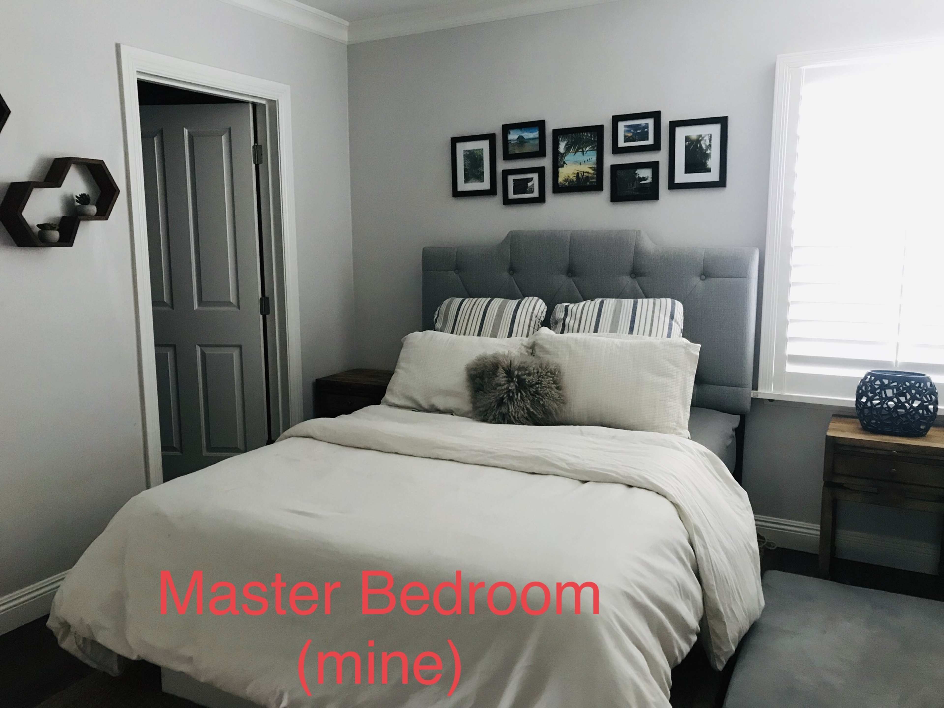 The image shows a master bedroom with a gray upholstered headboard, a neatly made bed with white bedding, and framed pictures on the wall.