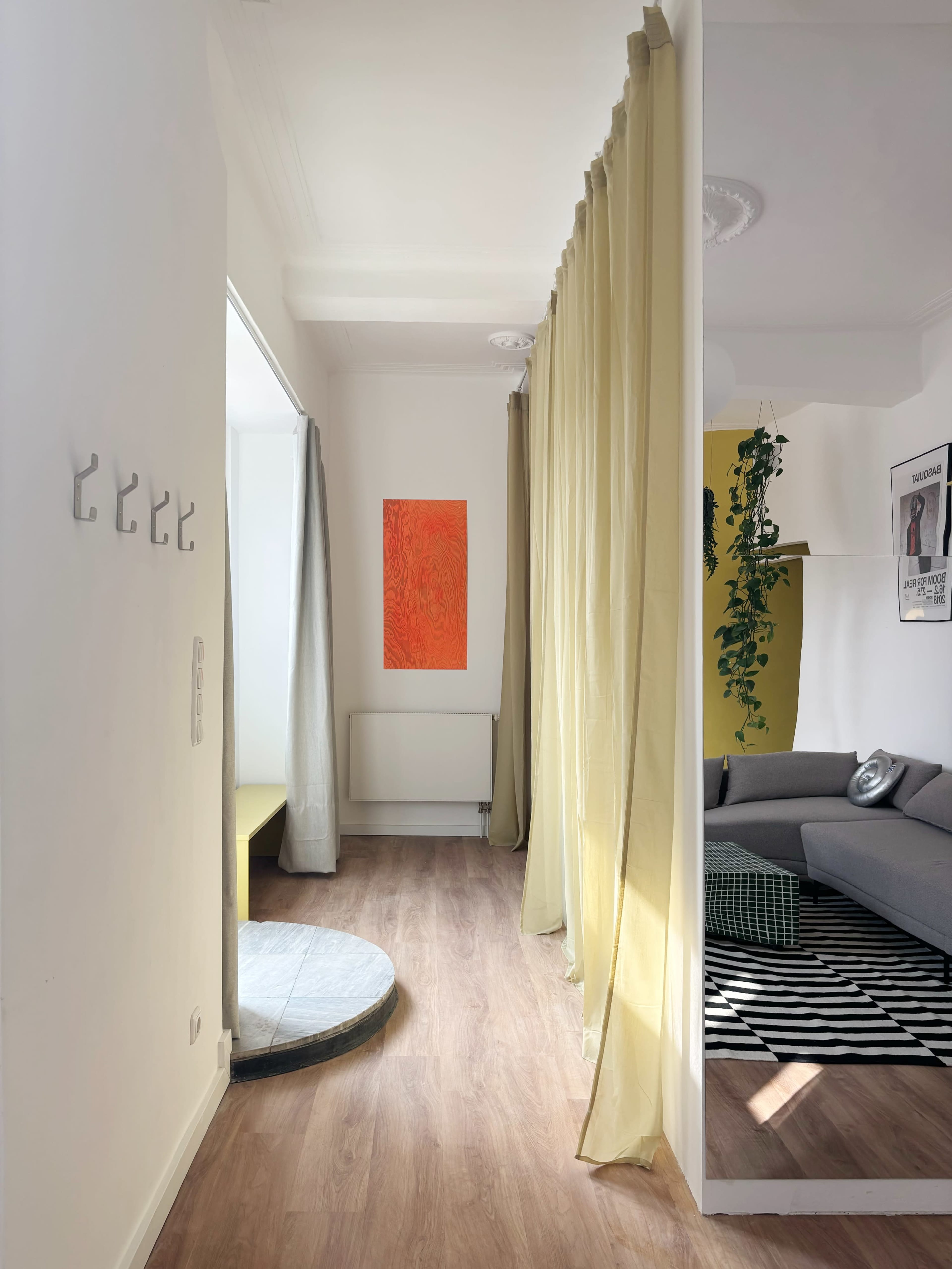 The image shows a bright room with a wooden floor, a gray sofa, a yellow wall, a colorful artwork, and a mirror reflecting the space.