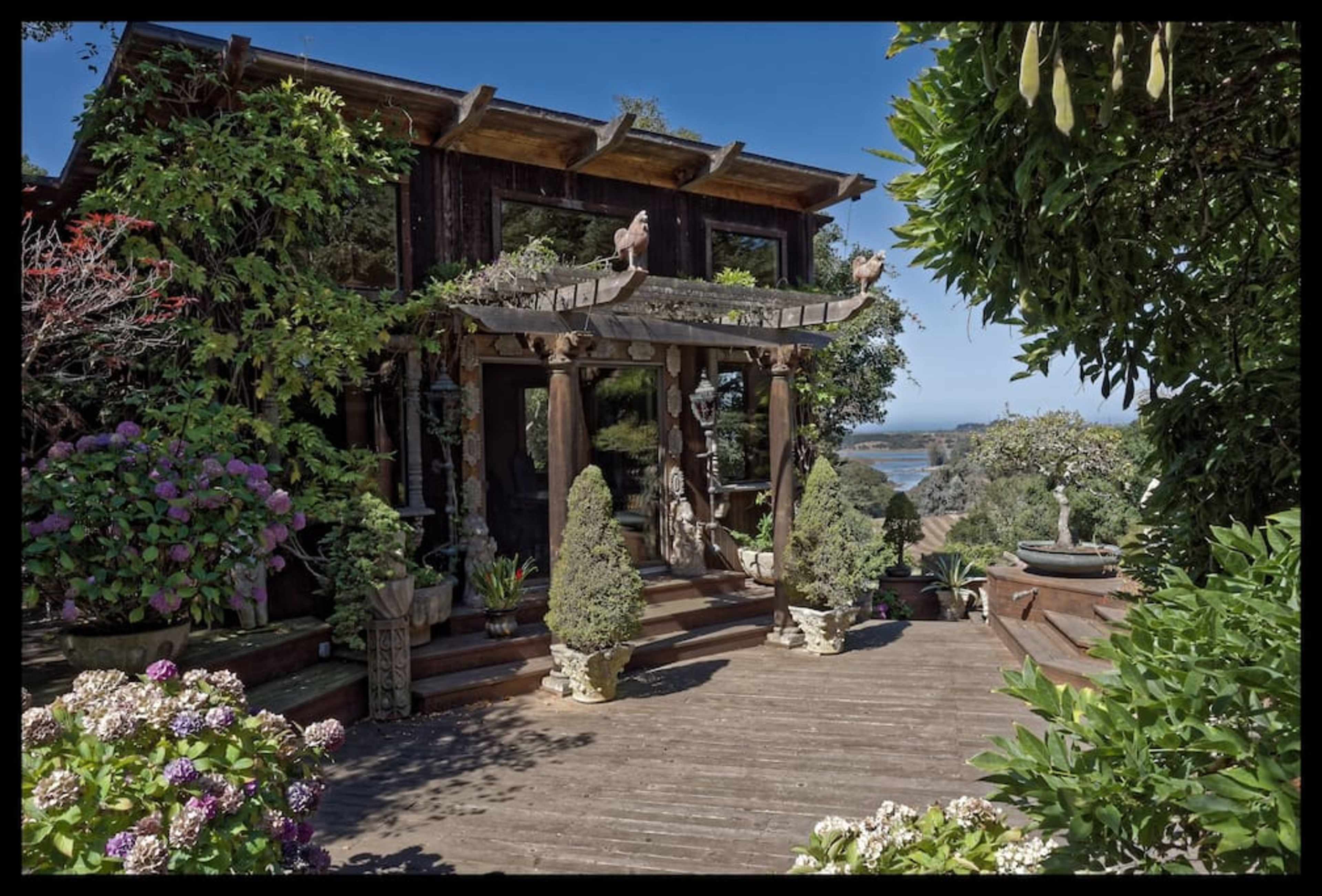 A wooden deck leads up to a house adorned with greenery and sculptures, overlooking a body of water.