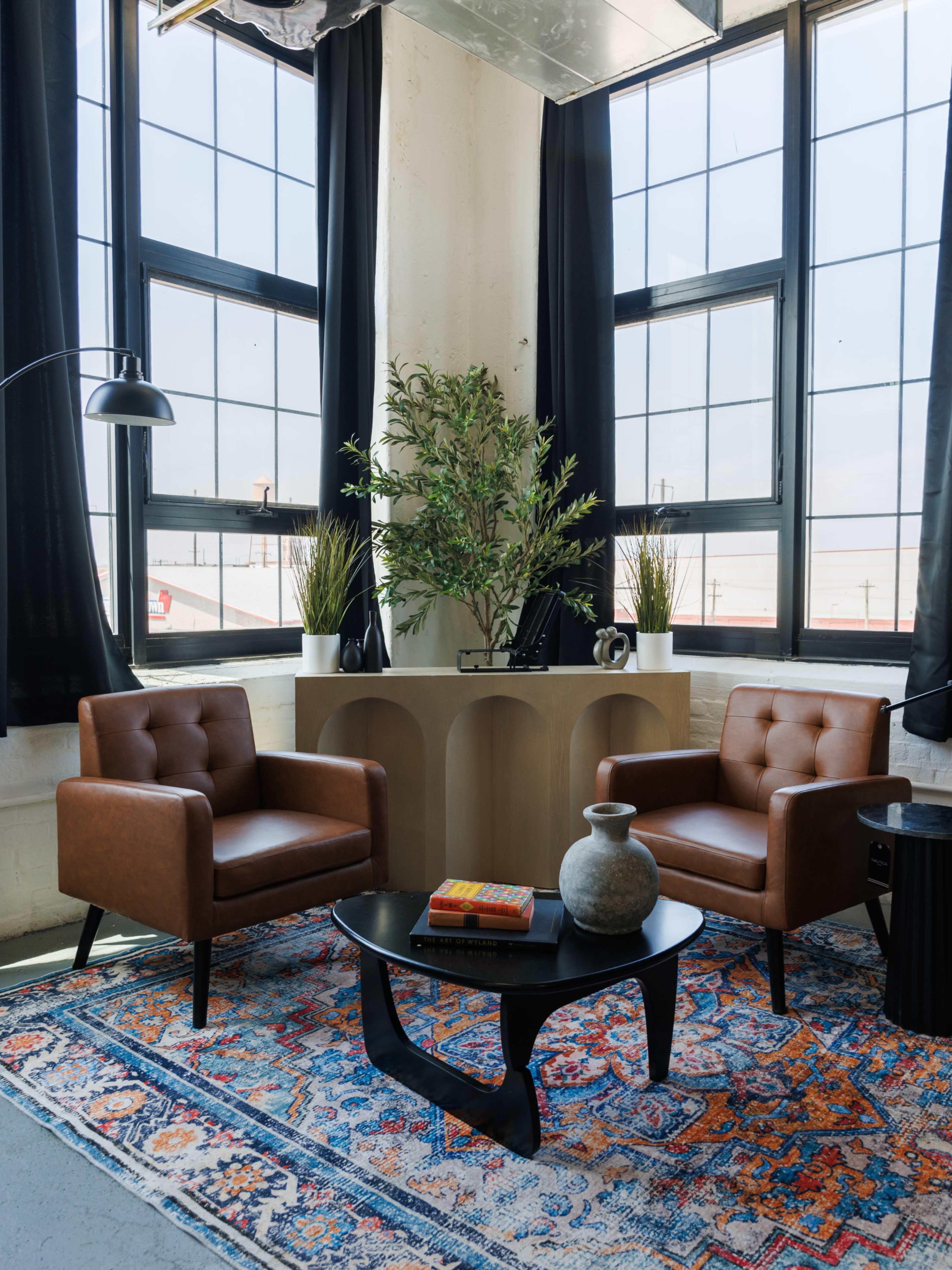 Two brown leather chairs are positioned across from a black coffee table in a well-lit room with large windows and a decorative plant.