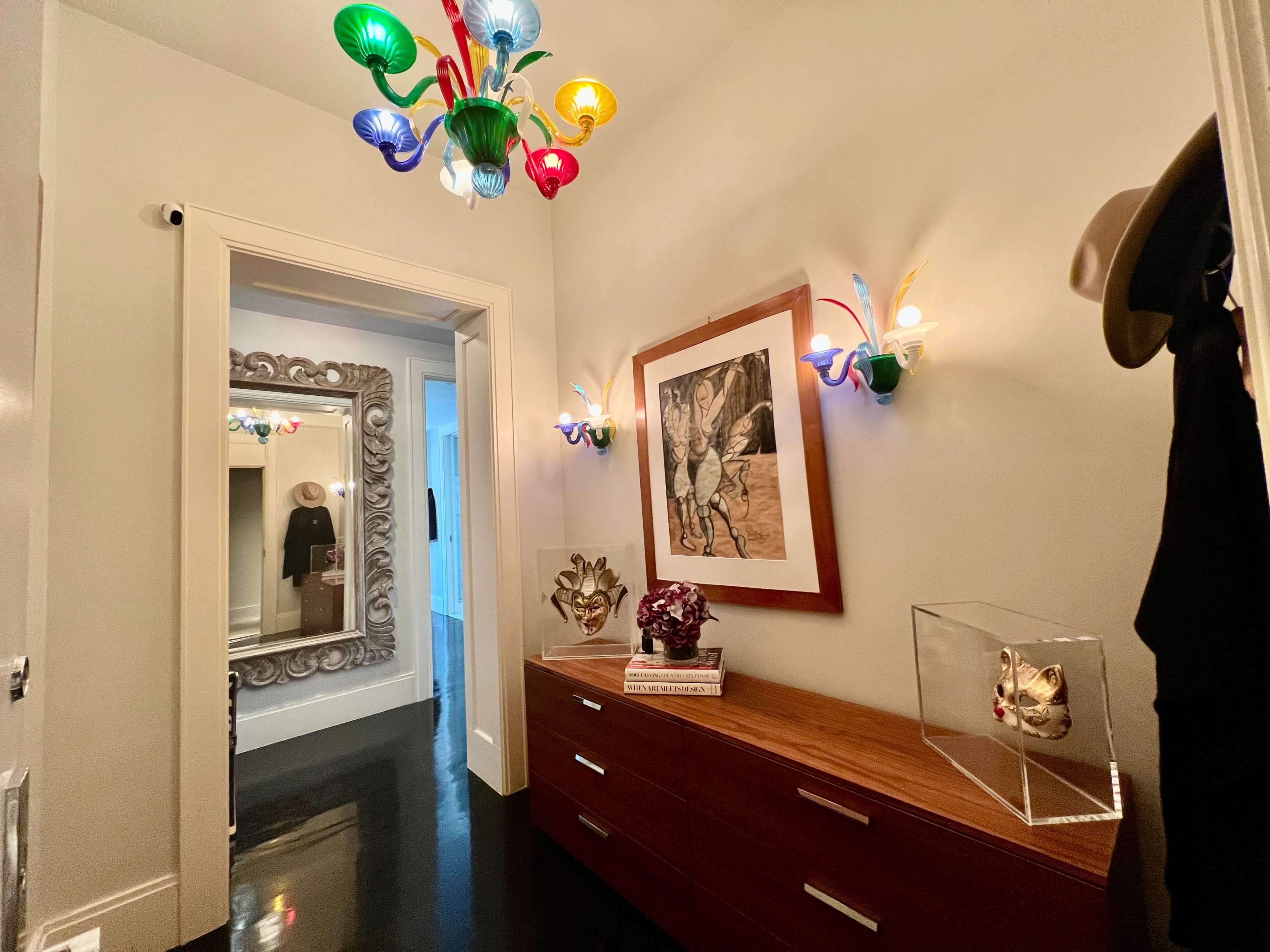 A hallway featuring a colorful chandelier, wall sconces, a large mirror with an ornate frame, and a wooden dresser with decorative items.