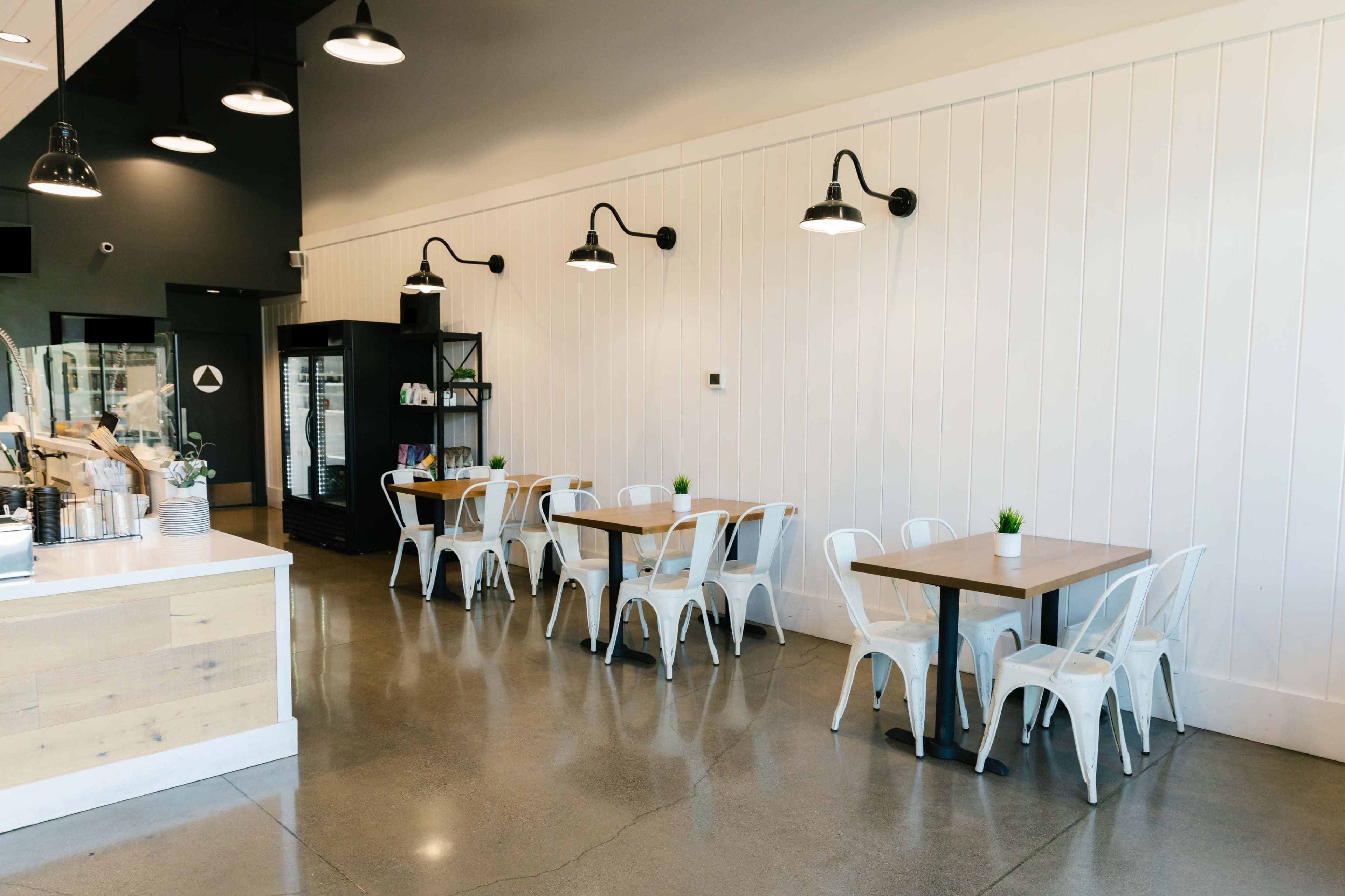 A modern cafe with white walls, metal chairs, and wooden tables arranged in a spacious layout, featuring overhead lights and a service counter.