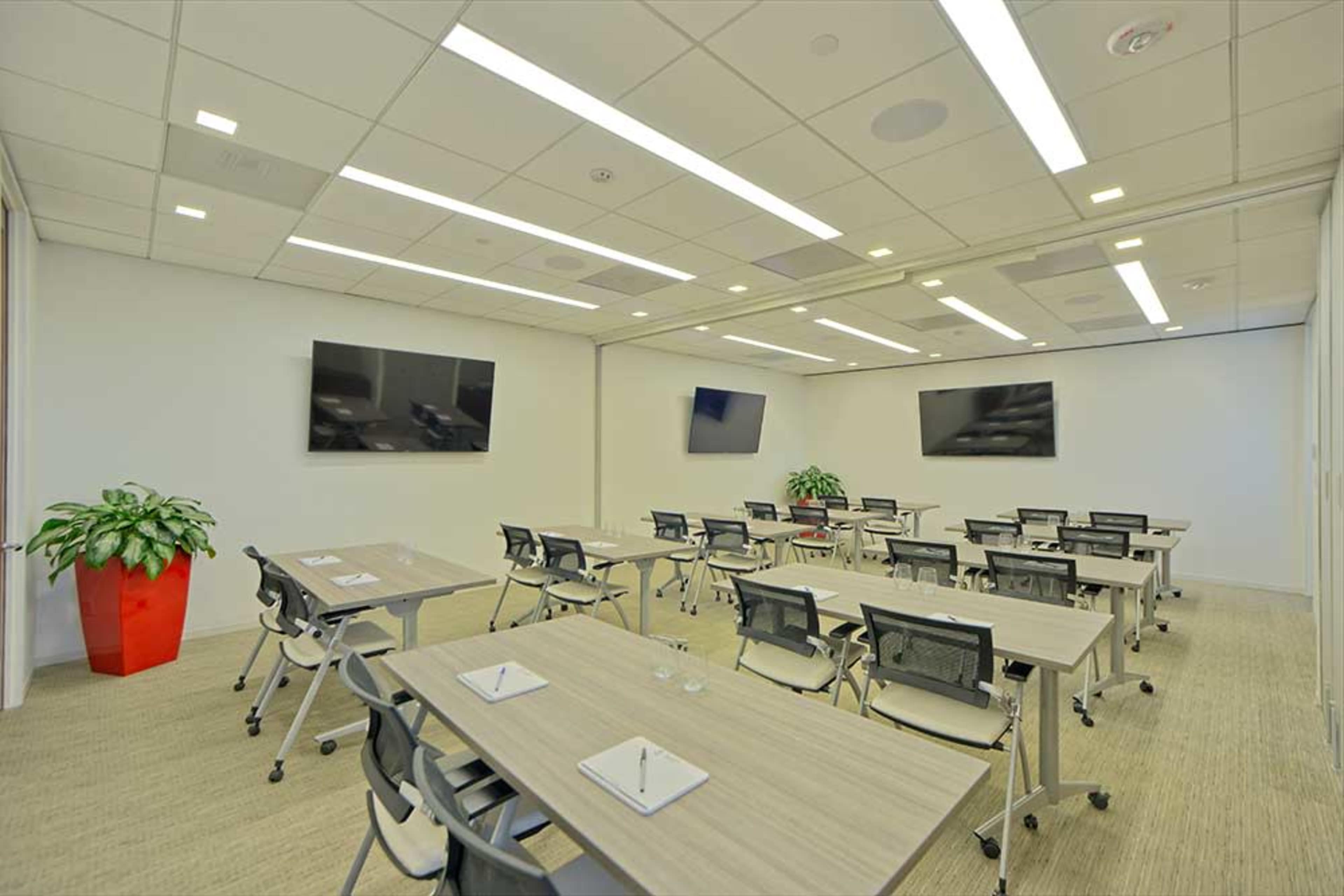 A modern conference room features multiple tables with chairs, two large screens mounted on the walls, and a potted plant in the corner.
