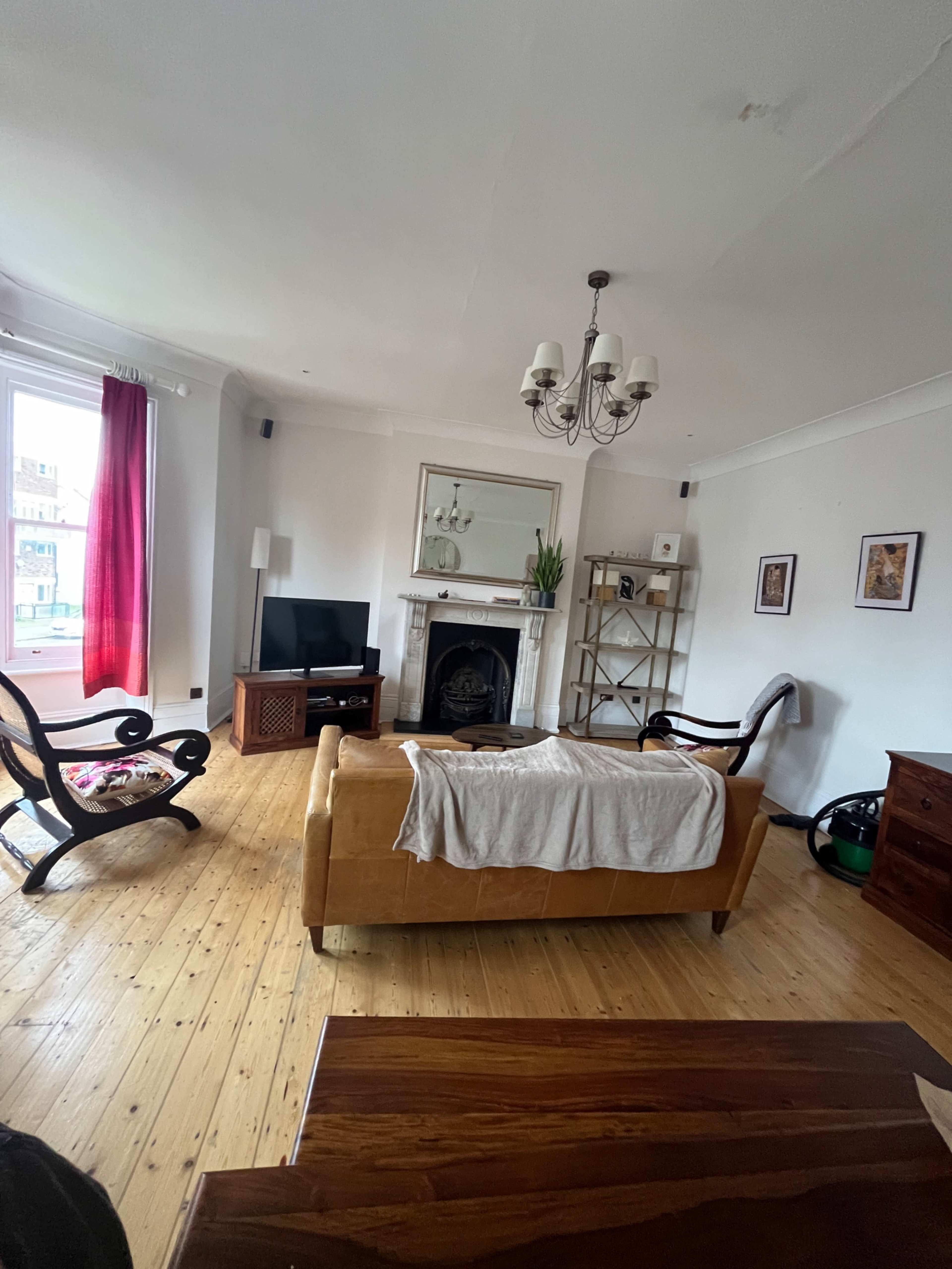 The room features a leather sofa, a wooden table, a television on a stand, a decorative fireplace, and a bookshelf, all set against a backdrop of light-colored walls and wooden flooring.