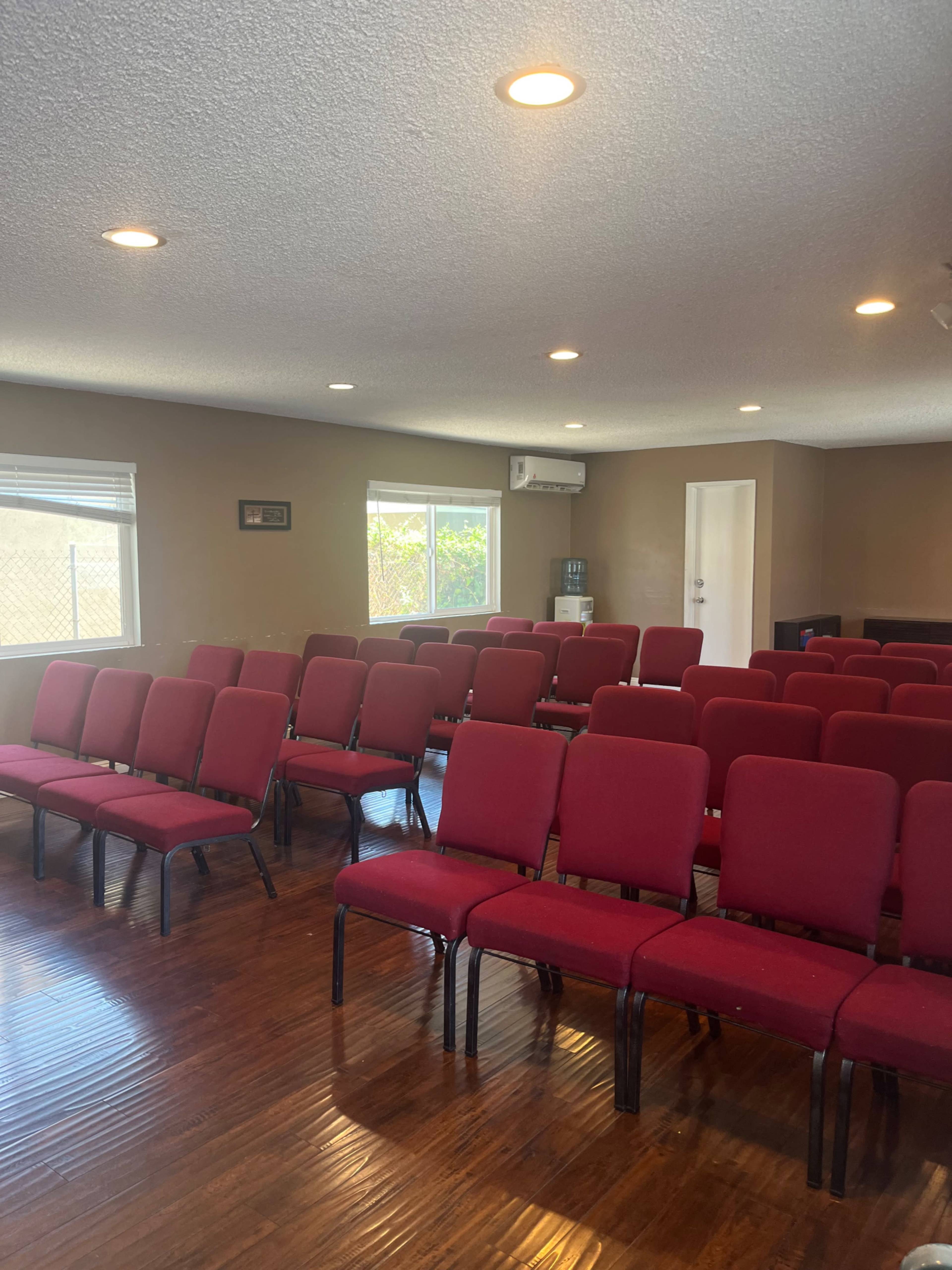 A room with rows of red chairs arranged facing the front, illuminated by ceiling lights and natural light from the windows.