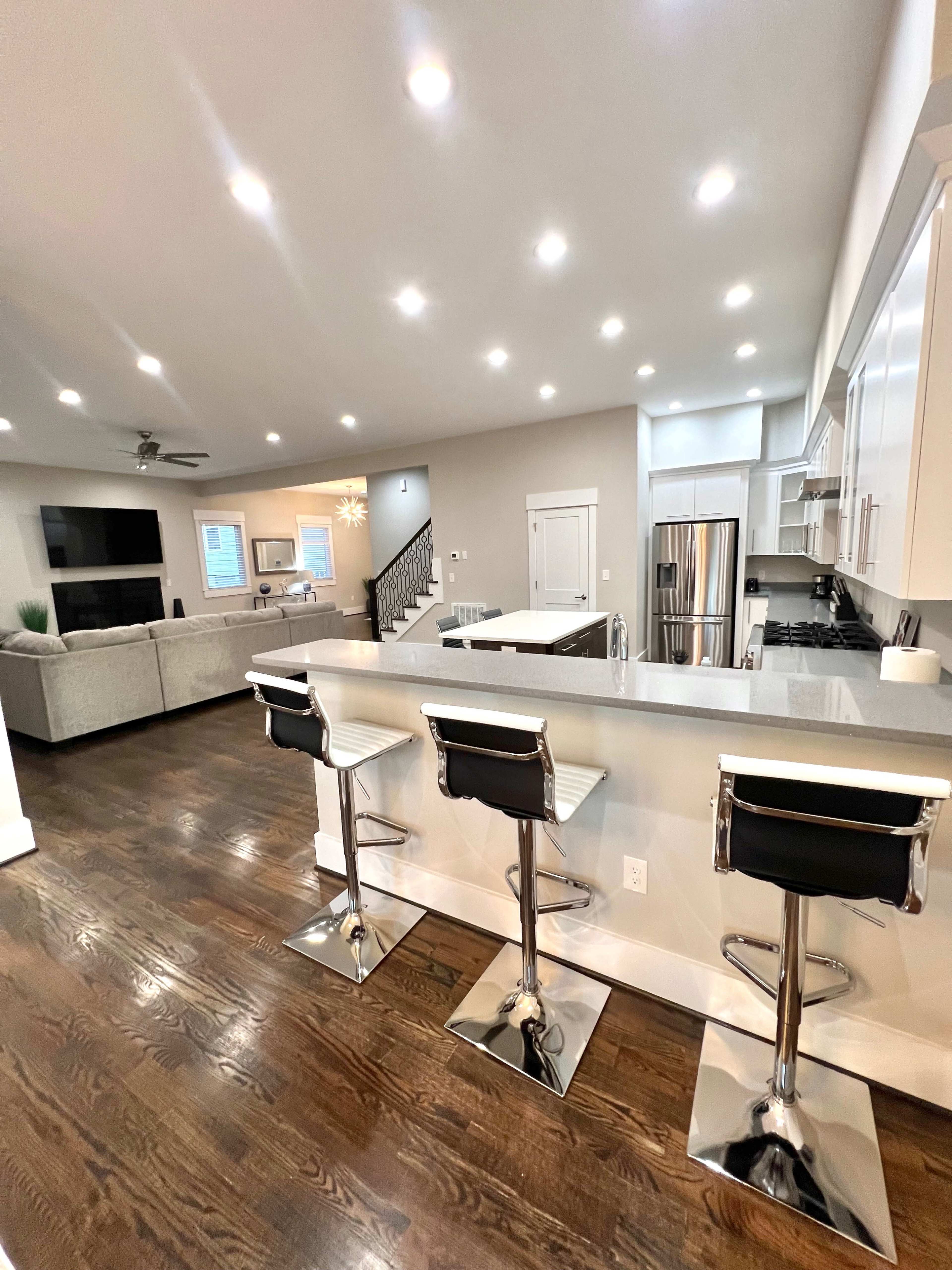 A modern kitchen features sleek bar stools at a counter, overlooking an open living space with a sofa and television.