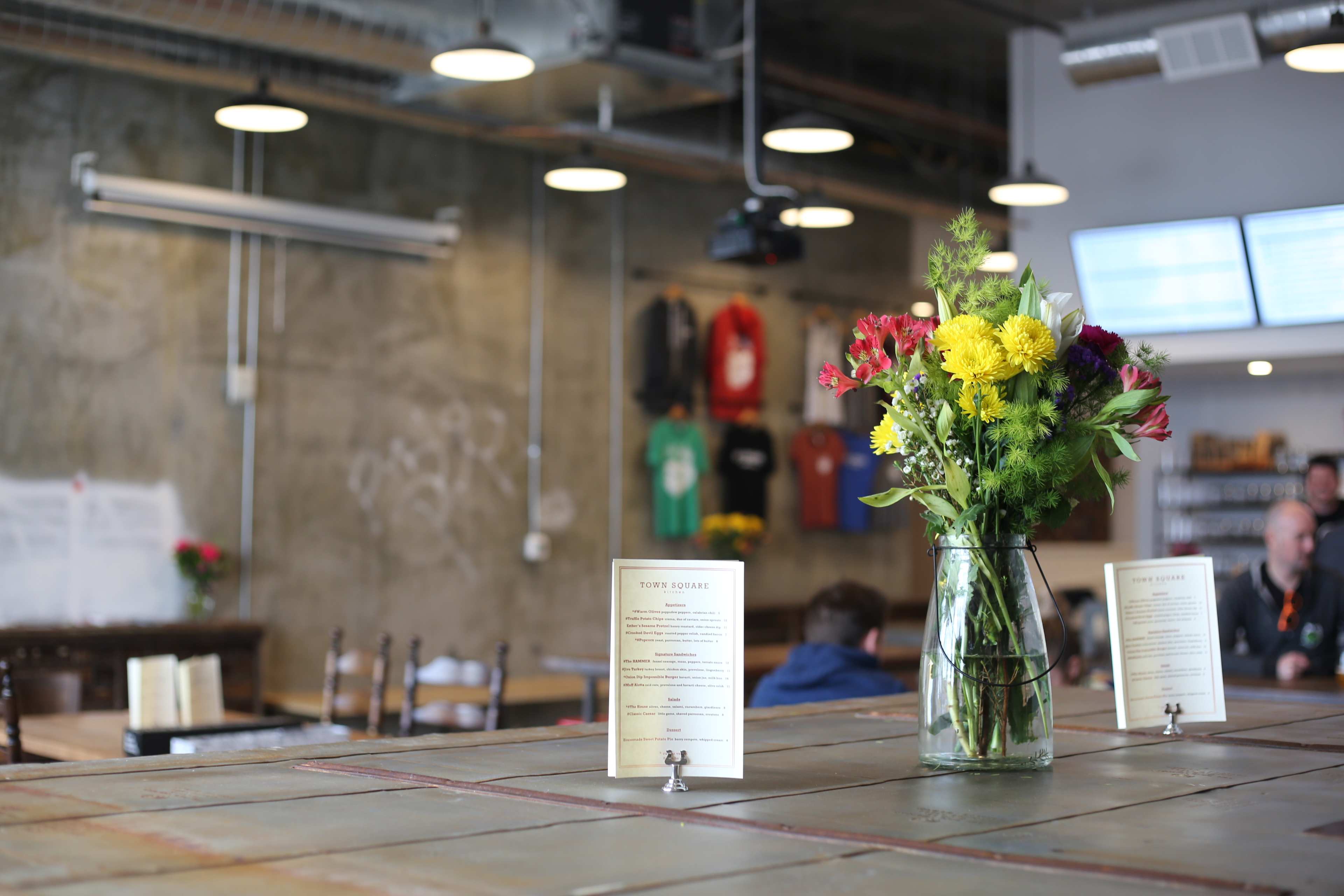 A flower arrangement in a glass vase sits on a table in a café featuring exposed concrete walls and menu cards.