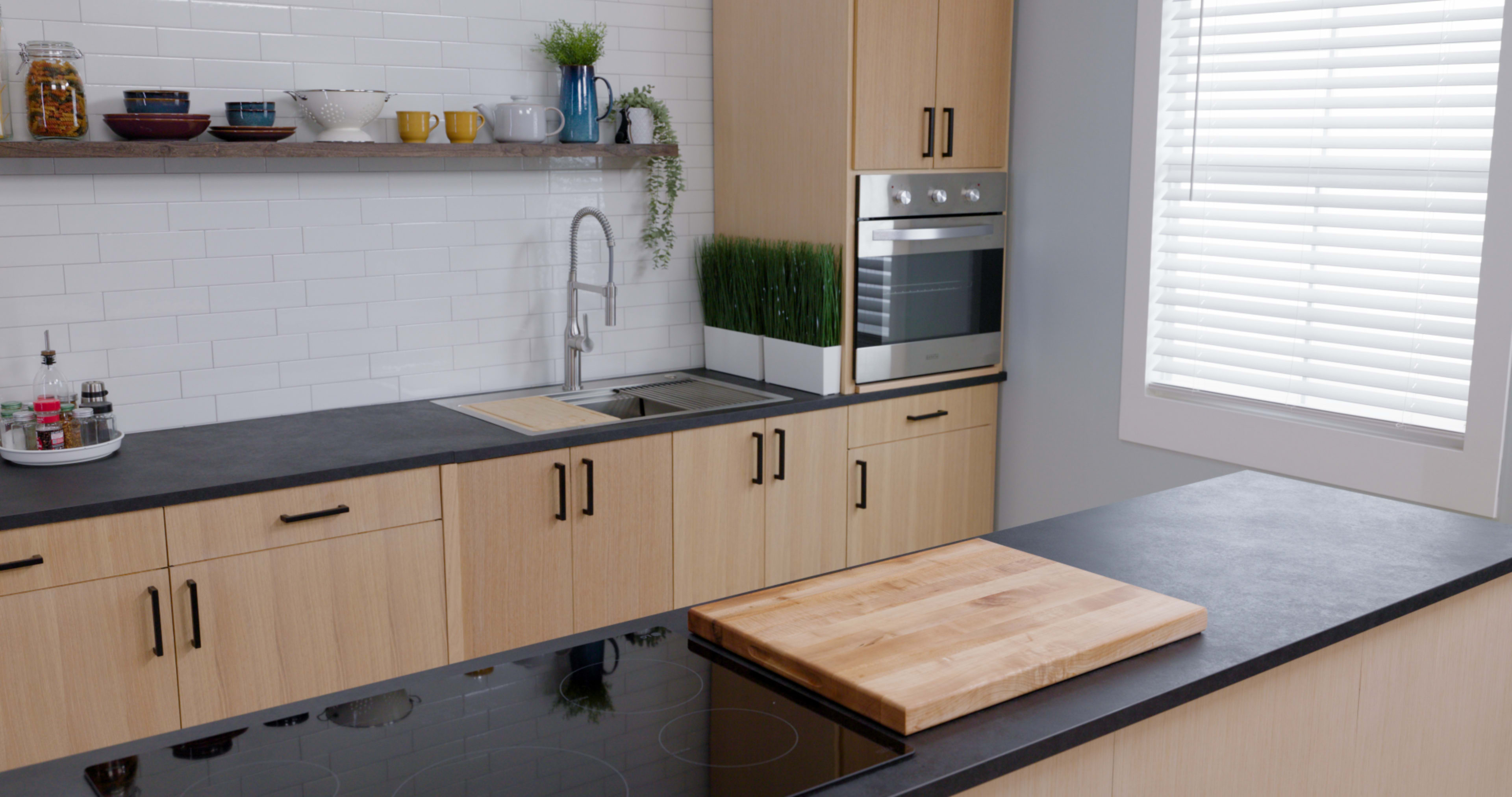 A modern kitchen features wooden cabinets, a sleek black countertop, a built-in oven, and a cutting board on a cooking surface.