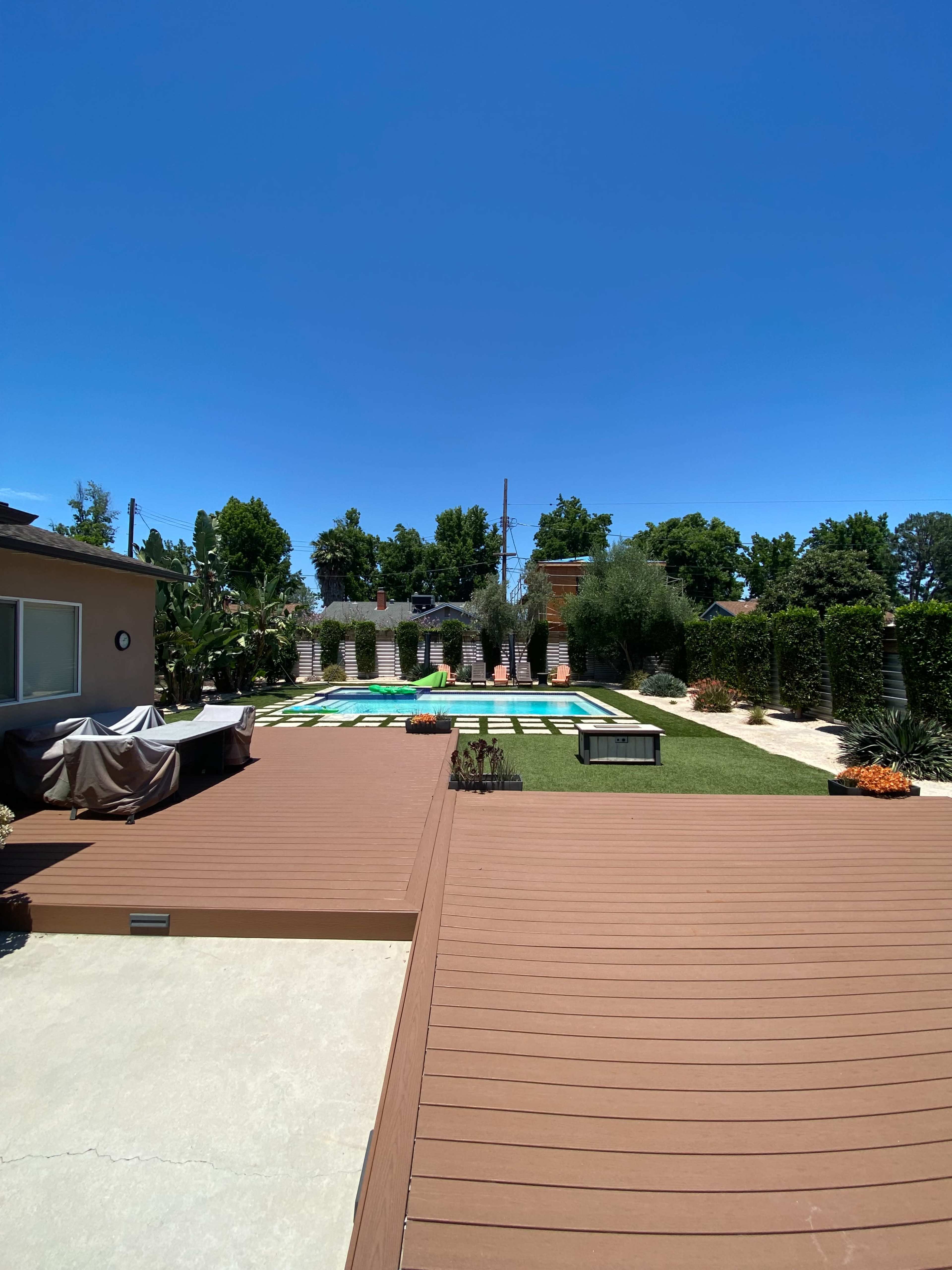 The image shows a backyard with a wooden deck, a swimming pool in the center, and landscaped greenery surrounding the area under a clear blue sky.