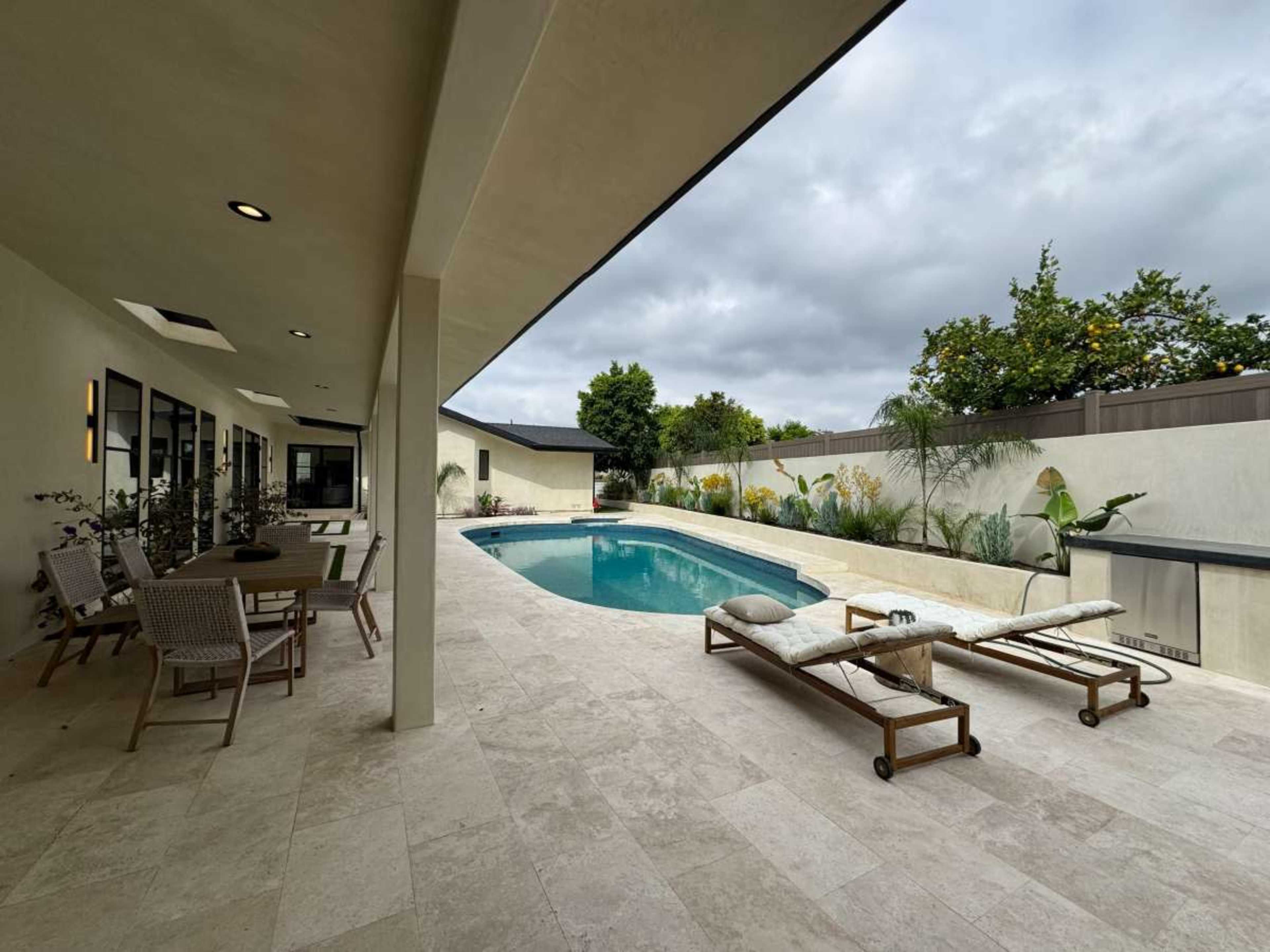 The image shows a modern backyard with a swimming pool, lounge chairs, and dining furniture, surrounded by greenery and low walls.