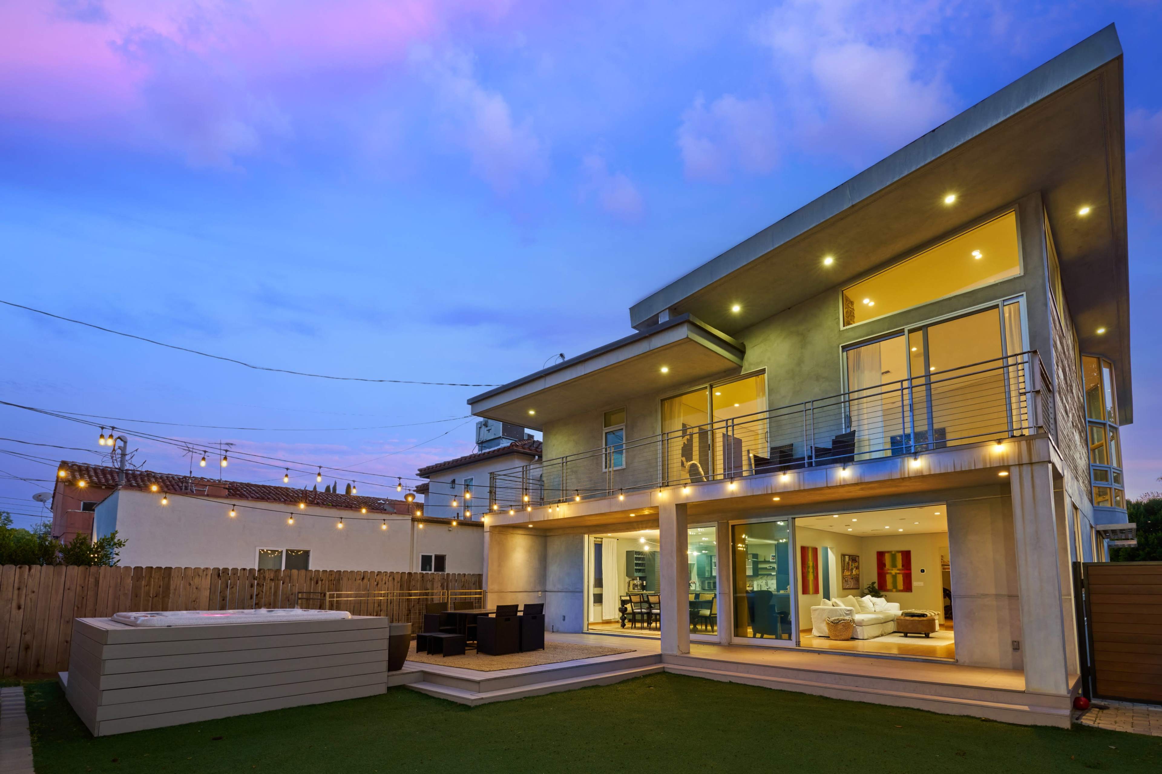 A modern two-story house is illuminated by string lights in the backyard, with large glass windows showcasing a living space and a hot tub on the patio.