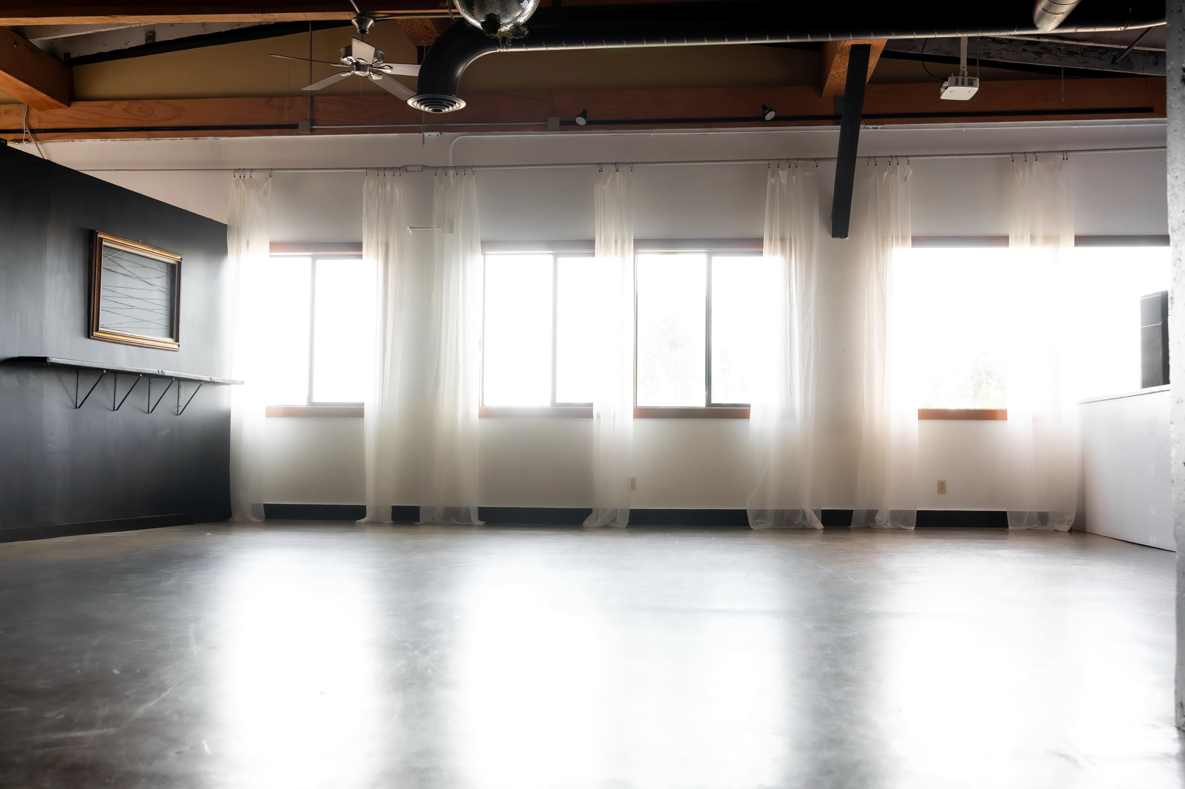 The image shows an empty room with large windows, sheer white curtains, a black wall with a framed board, and polished concrete flooring.