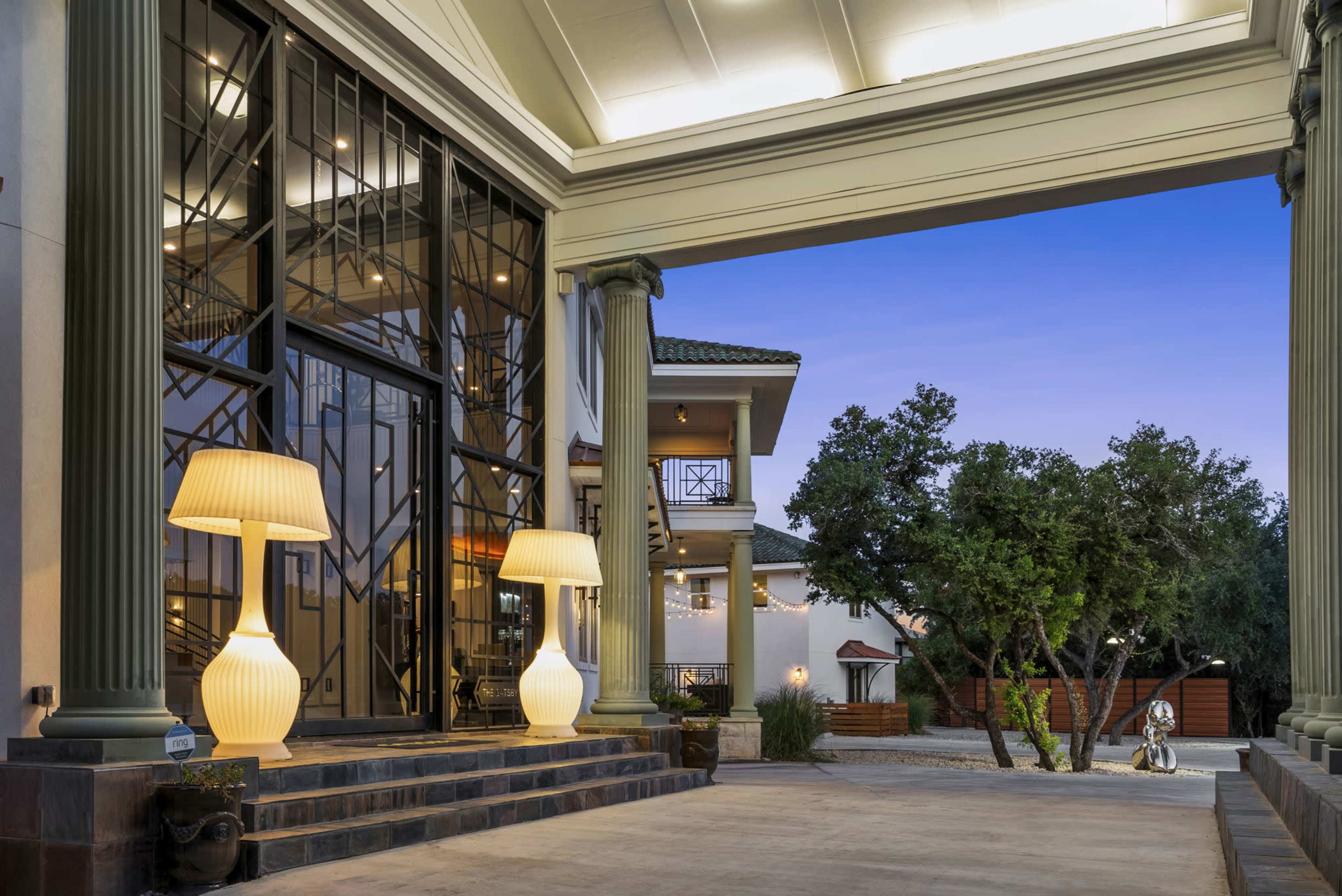 A large entrance with stylish columned architecture, featuring two illuminated lamps, and a view of a landscaped area with modern sculptures in the background.
